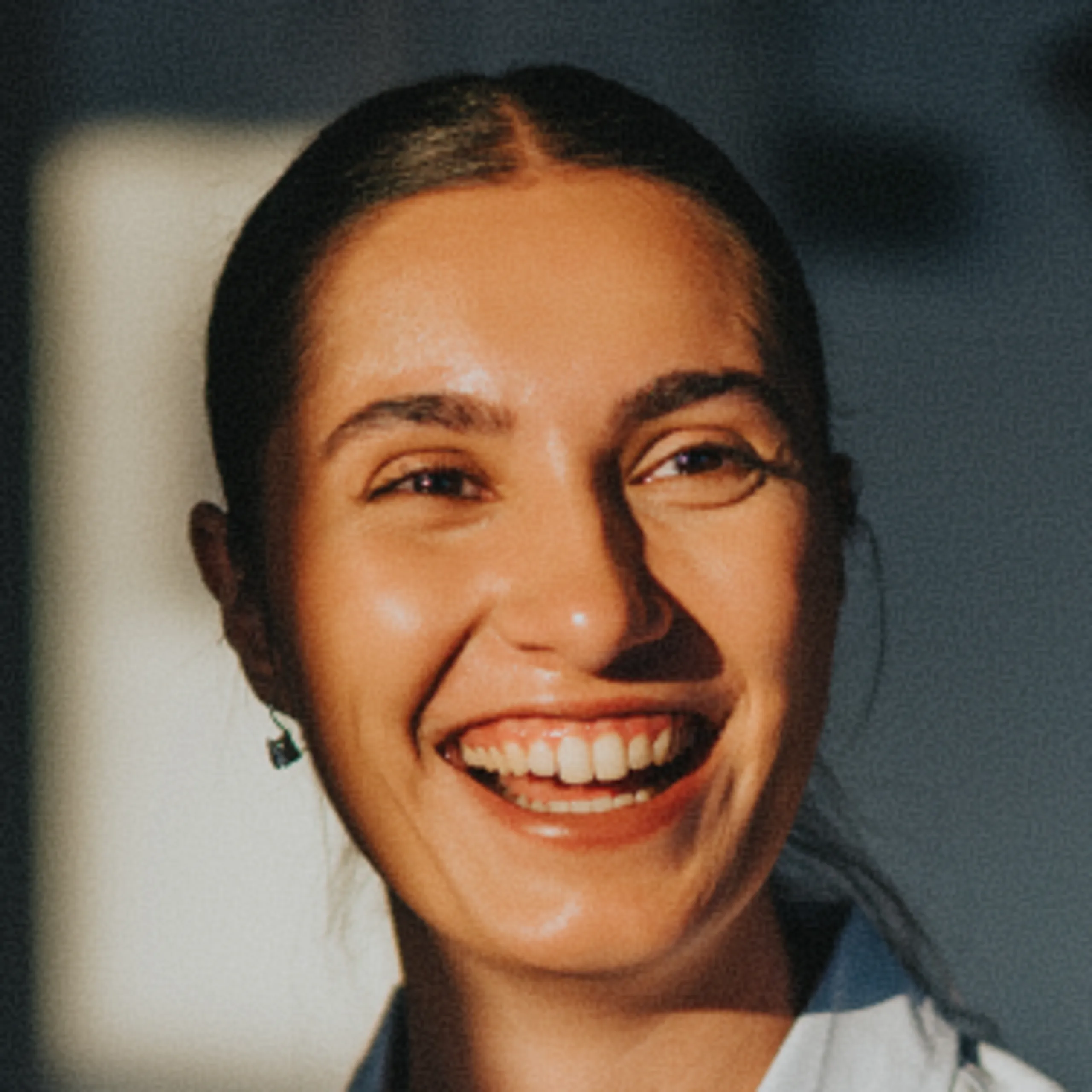 A woman with dark hair pulled back smiles brightly in warm sunlight, wearing a light-colored top and small earrings.
