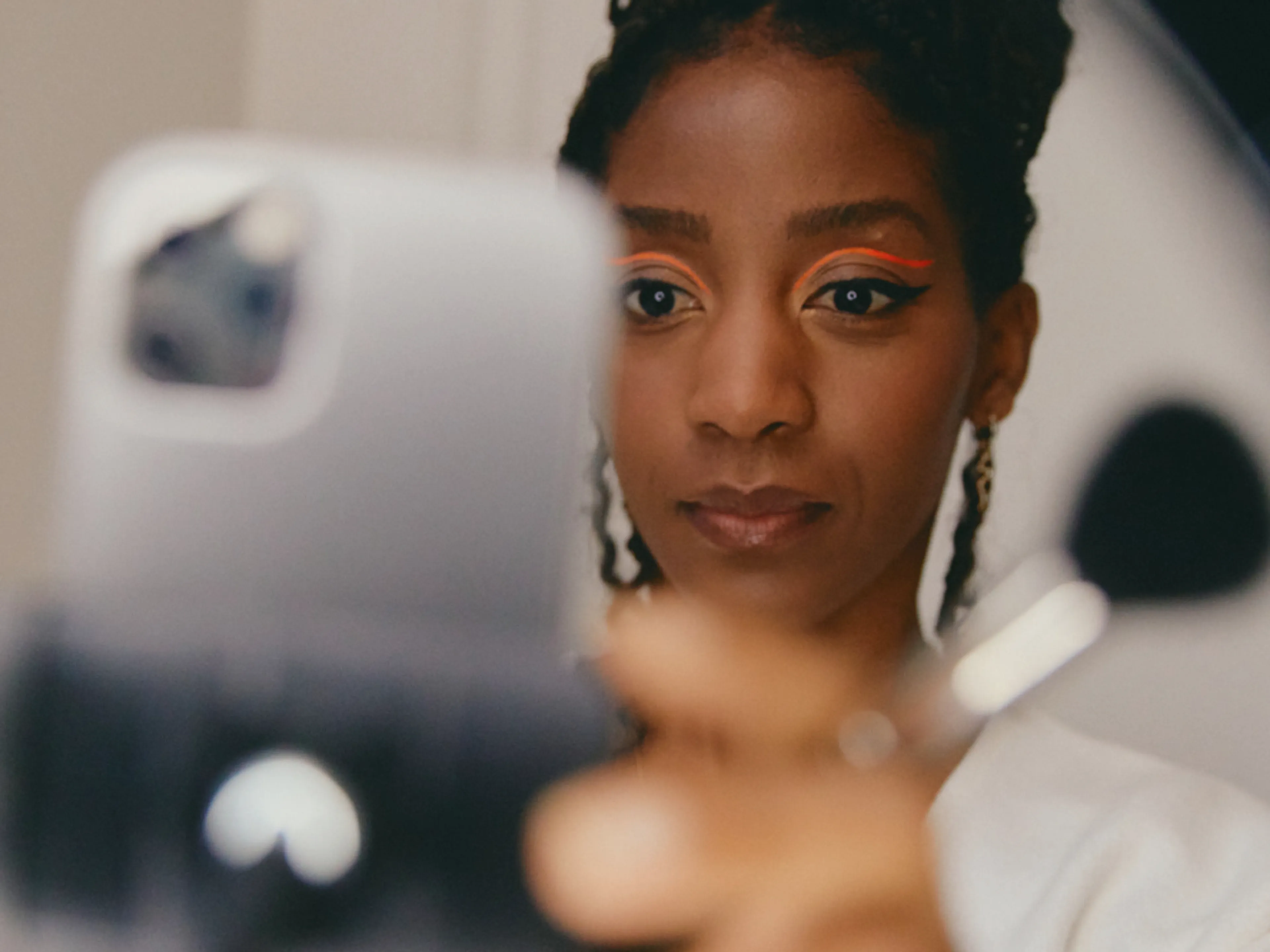 A woman with bold orange eyeliner takes a selfie with a smartphone, holding a makeup brush in her hand.