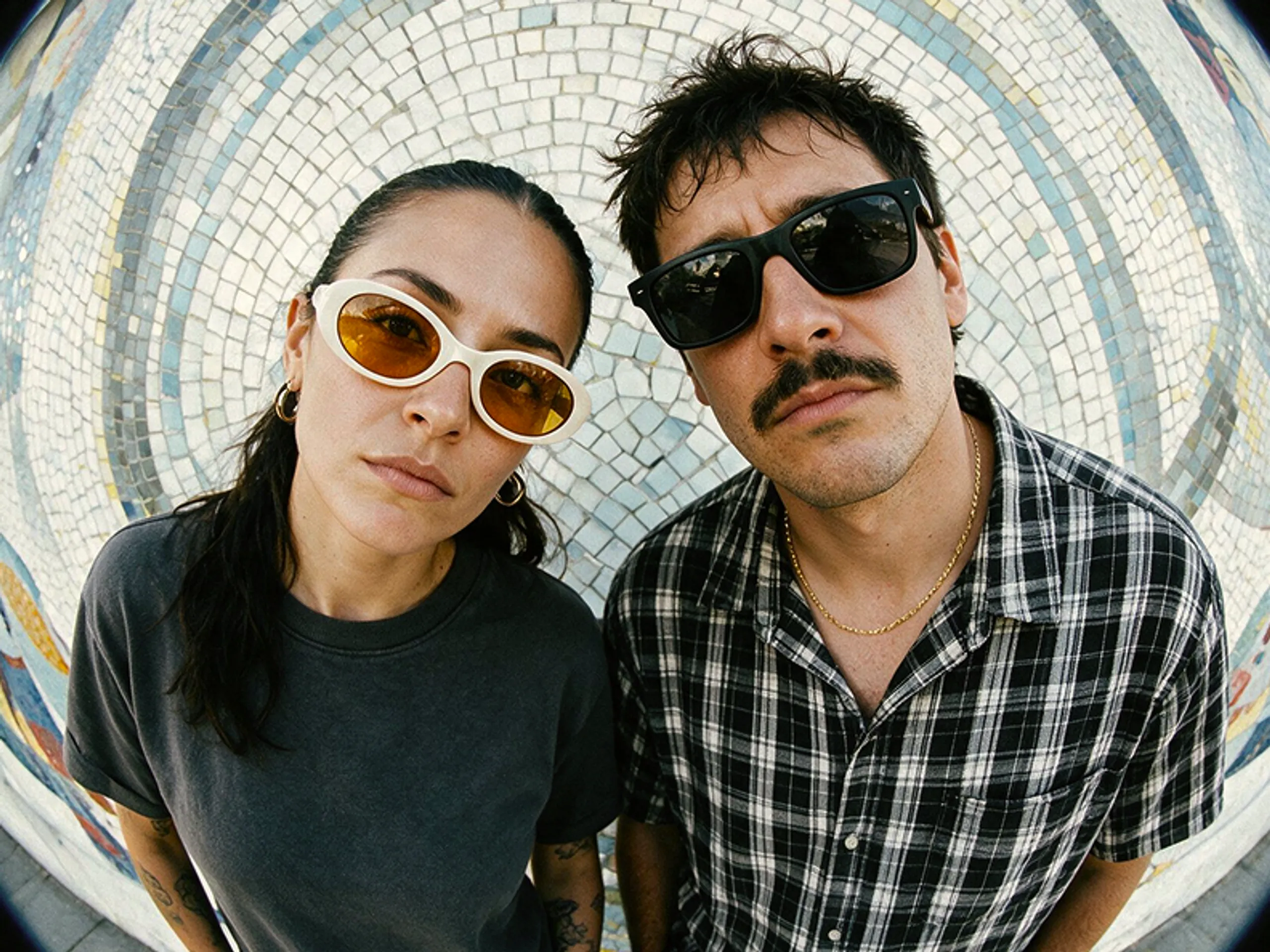 Two people wearing sunglasses and casual clothes stand close together, looking up at the camera with serious expressions. The background is a colorful, tiled mosaic captured through a fisheye lens.