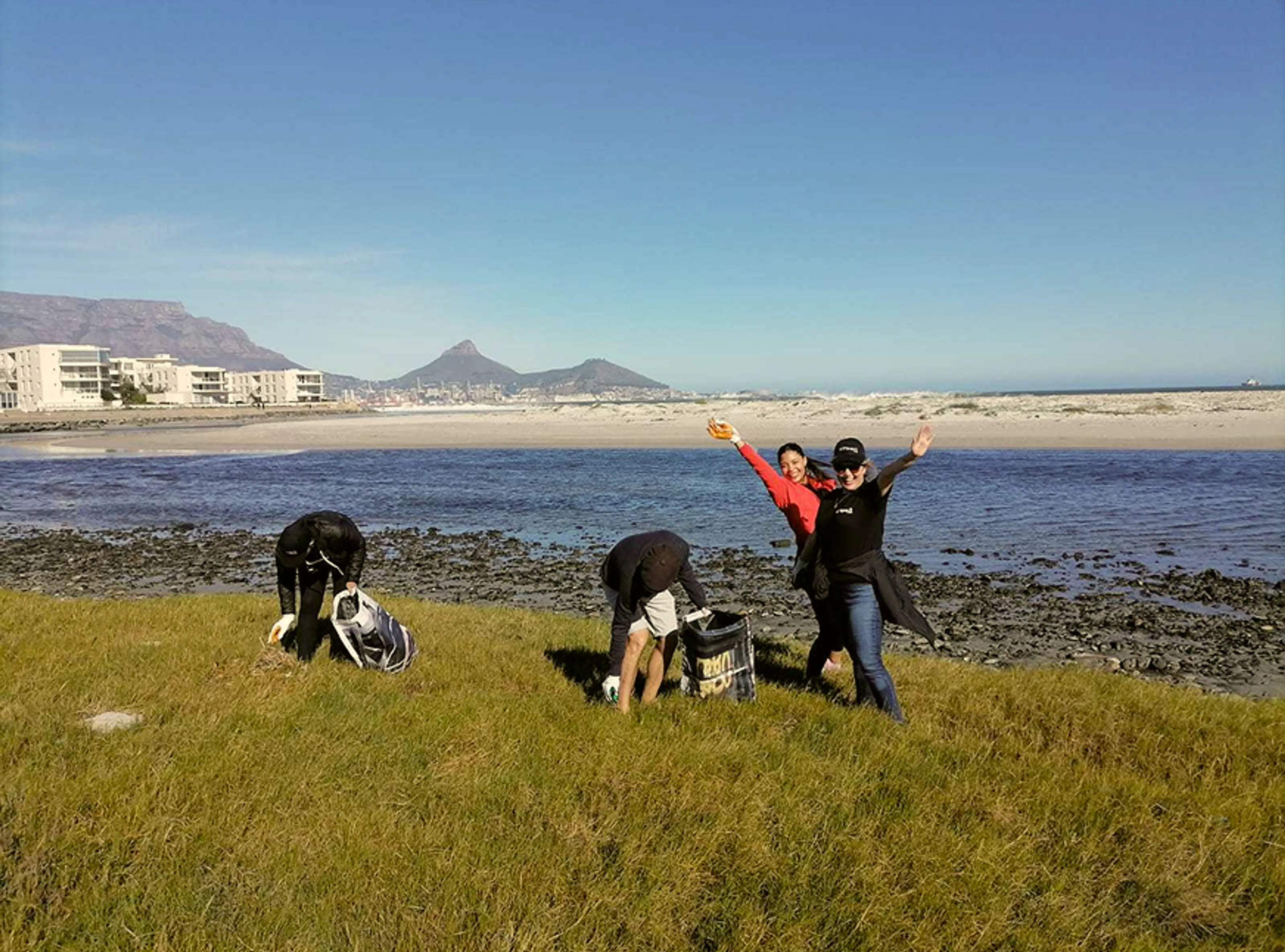 Four people clean up litter on a grassy shore near the beach. Two are bent over picking up trash, while two others stand, smiling and posing with arms raised. Ocean, buildings, and mountains are visible in the background.