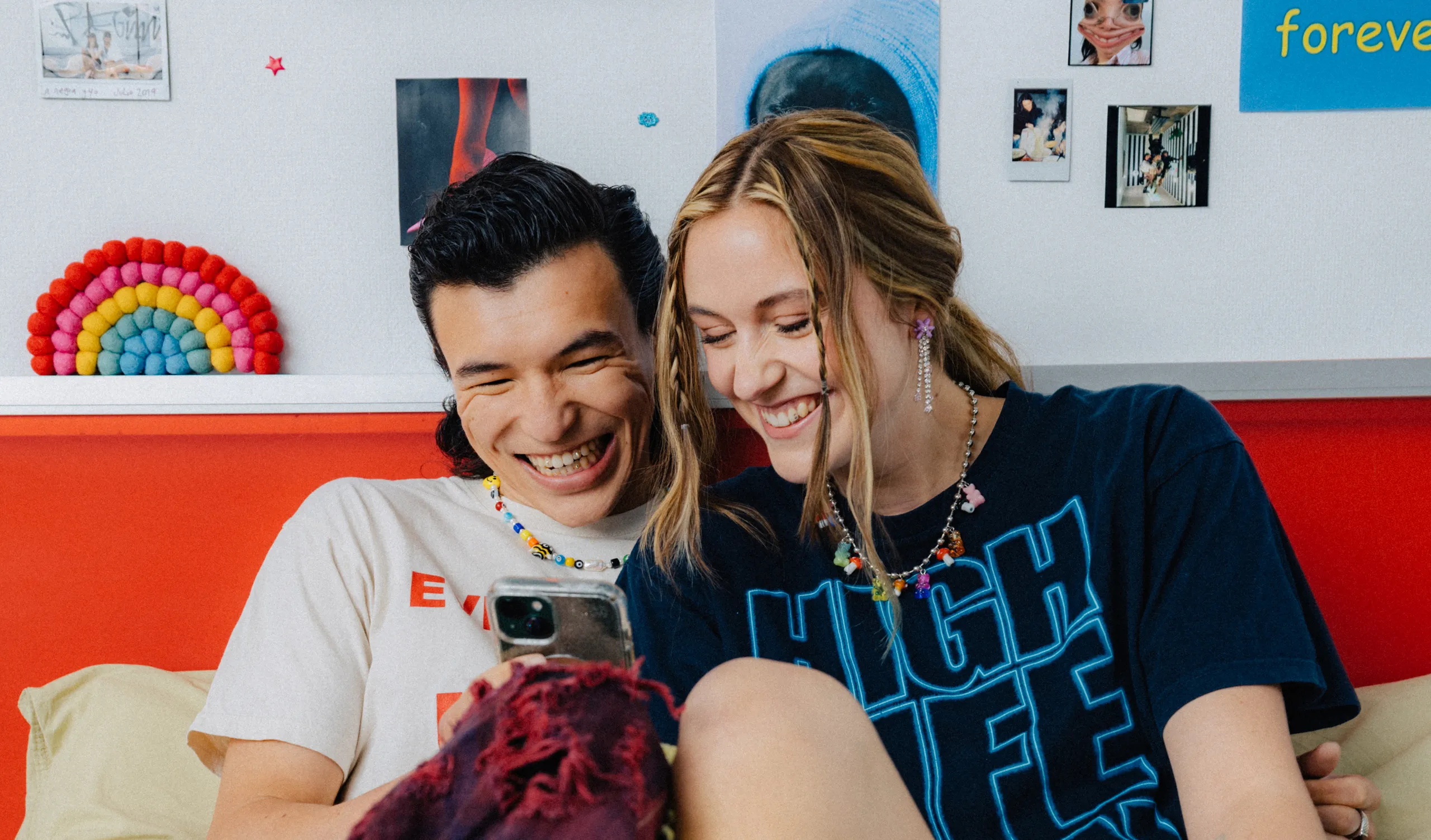 Two people sit closely on a bed, smiling and looking at a phone. The background has photos, a rainbow decoration, and colorful decor on the wall.
