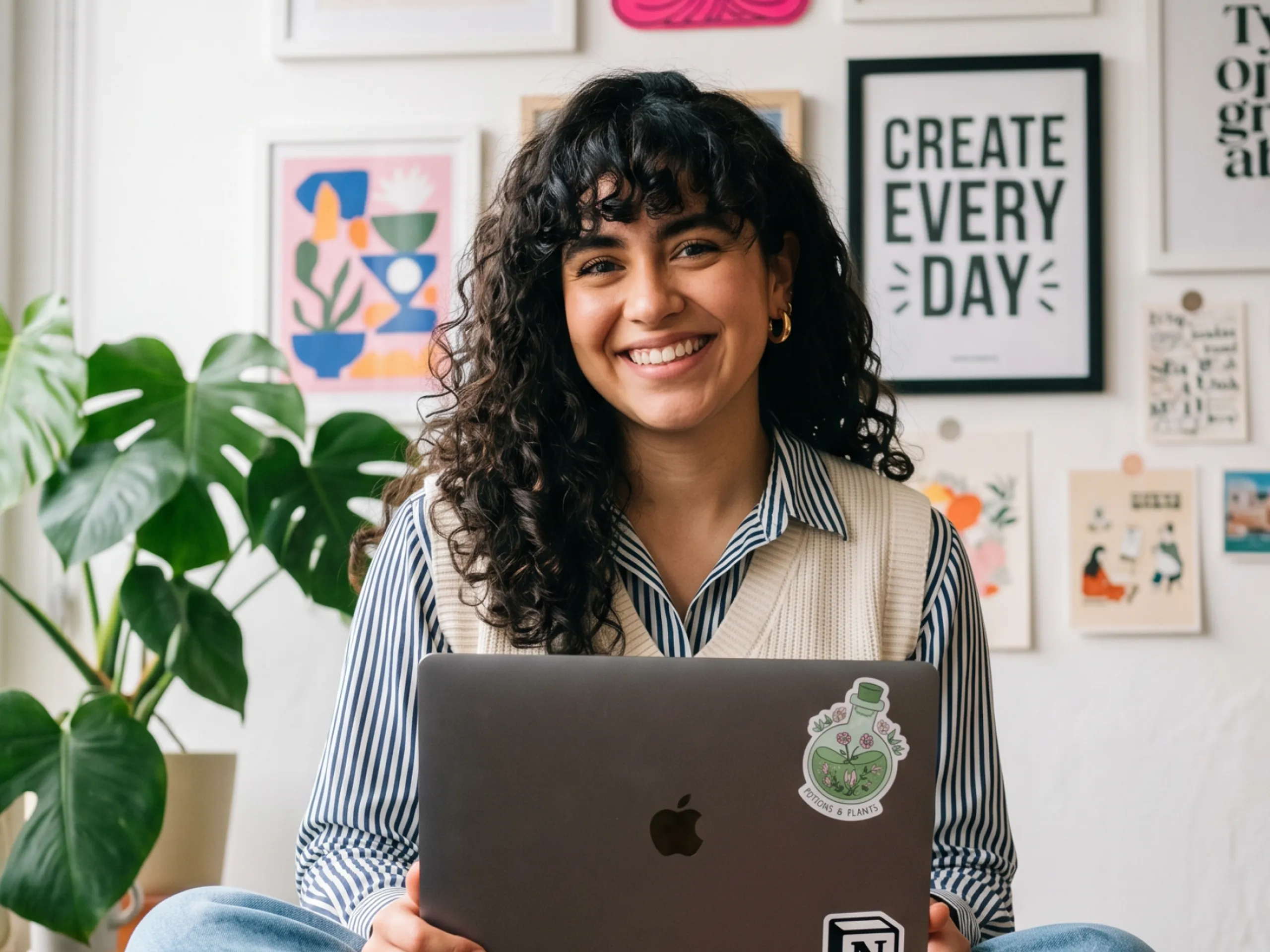 A smiling person with curly hair sits holding a laptop, in front of a wall with colorful art and motivational prints. A green plant is on the left side of the image.