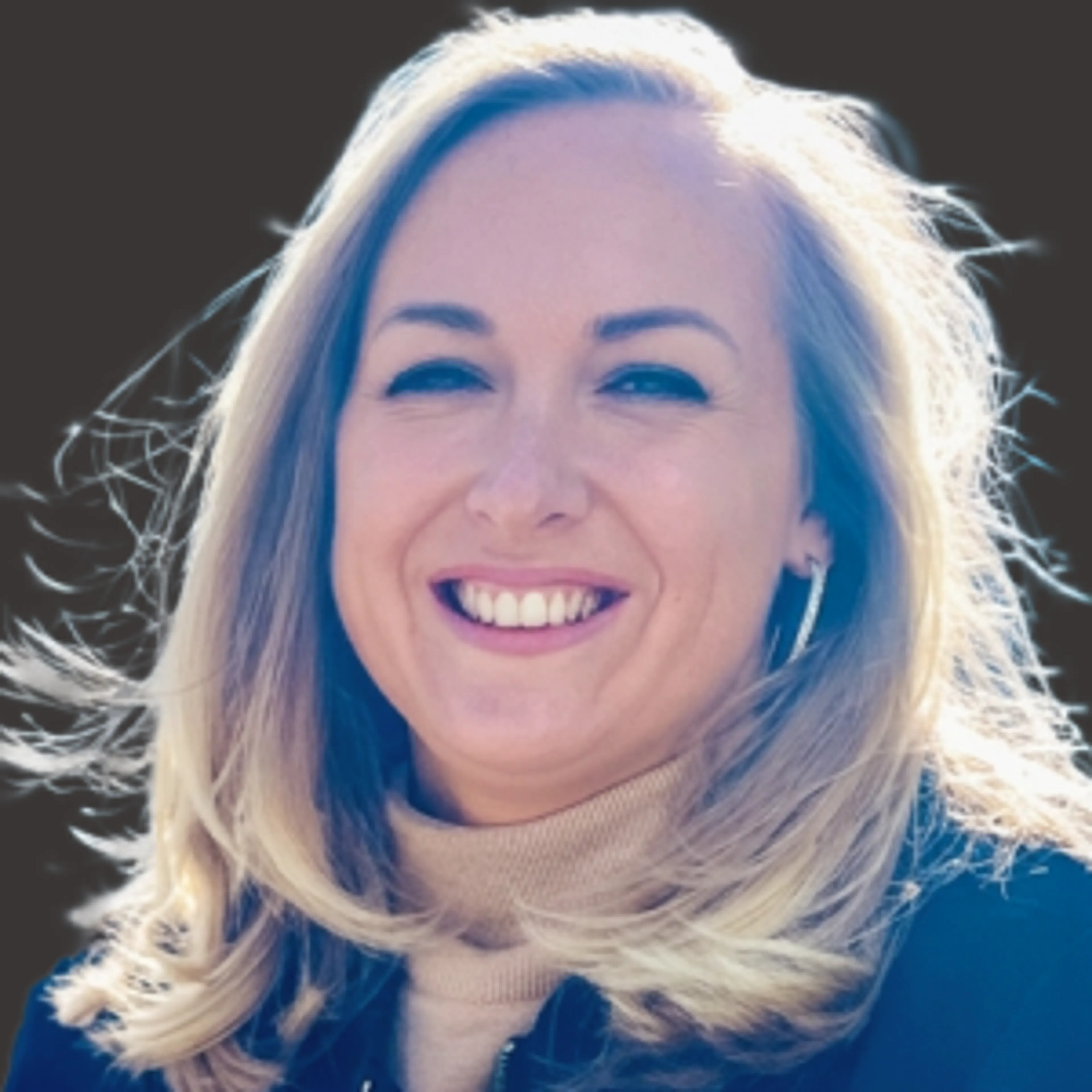 A smiling woman with blonde hair wearing a beige turtleneck and dark jacket, standing outdoors with wind blowing her hair and a dark background.