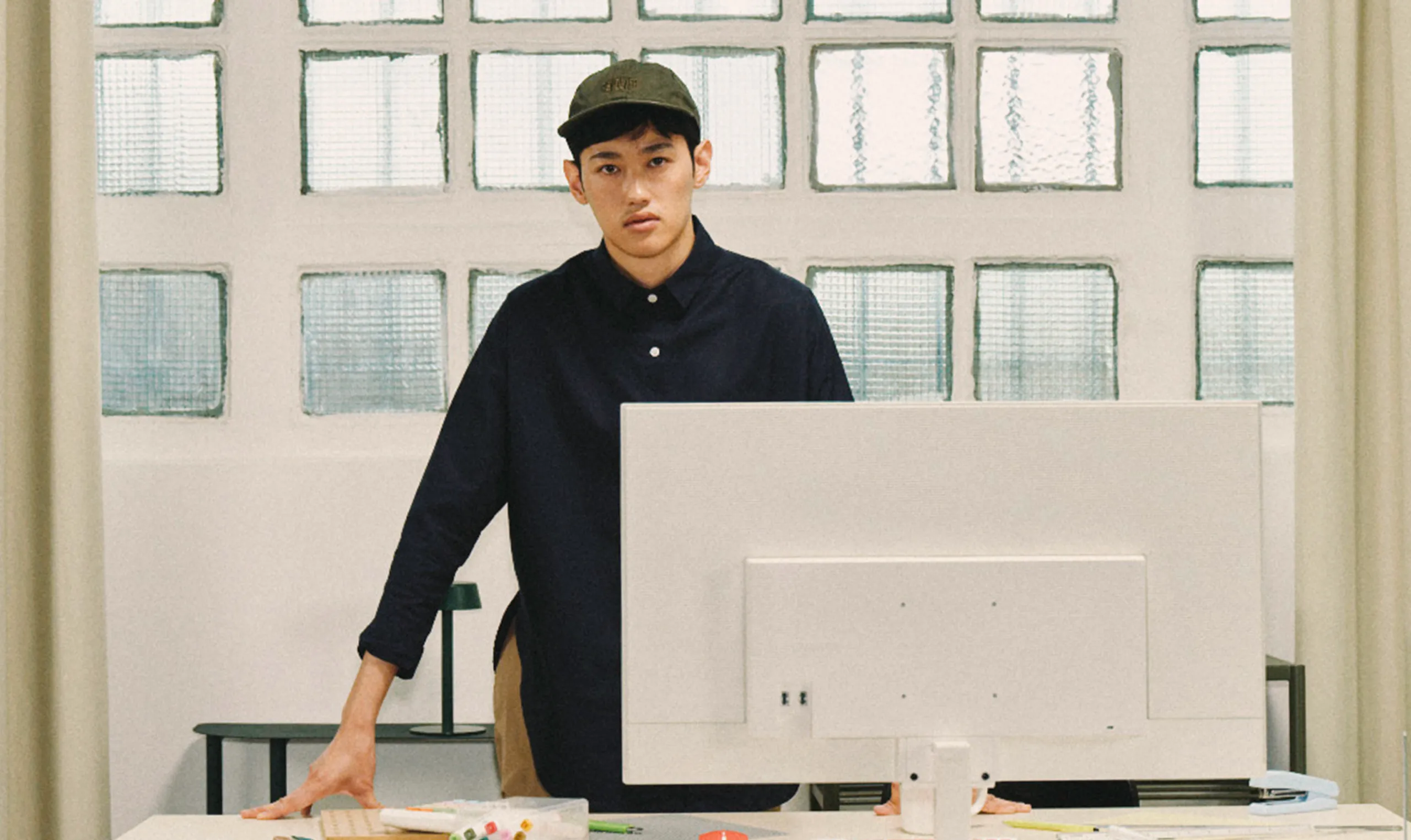 A young man in a dark shirt and cap stands behind a large white computer monitor in a modern office with glass block windows. His hands rest on the desk, and he faces the camera with a neutral expression.