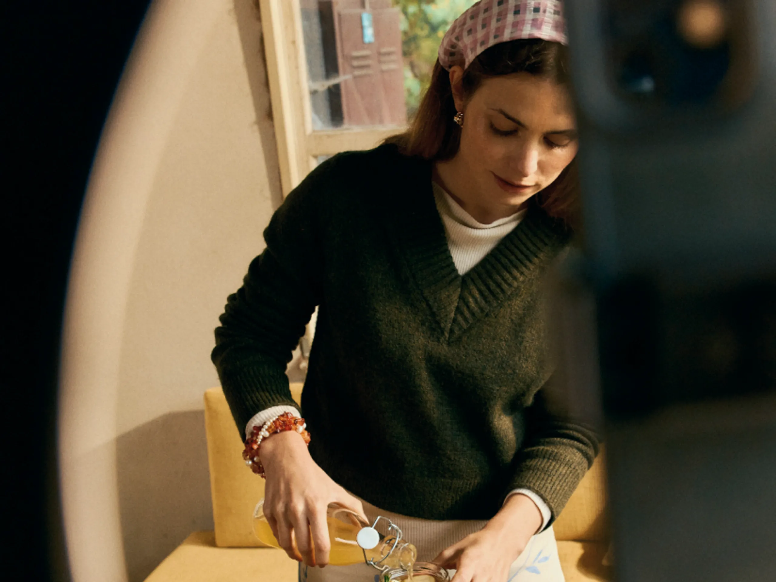 A woman wearing a dark green sweater and a pink headband pours a drink into a glass at a table indoors, with soft light coming from a nearby window.
