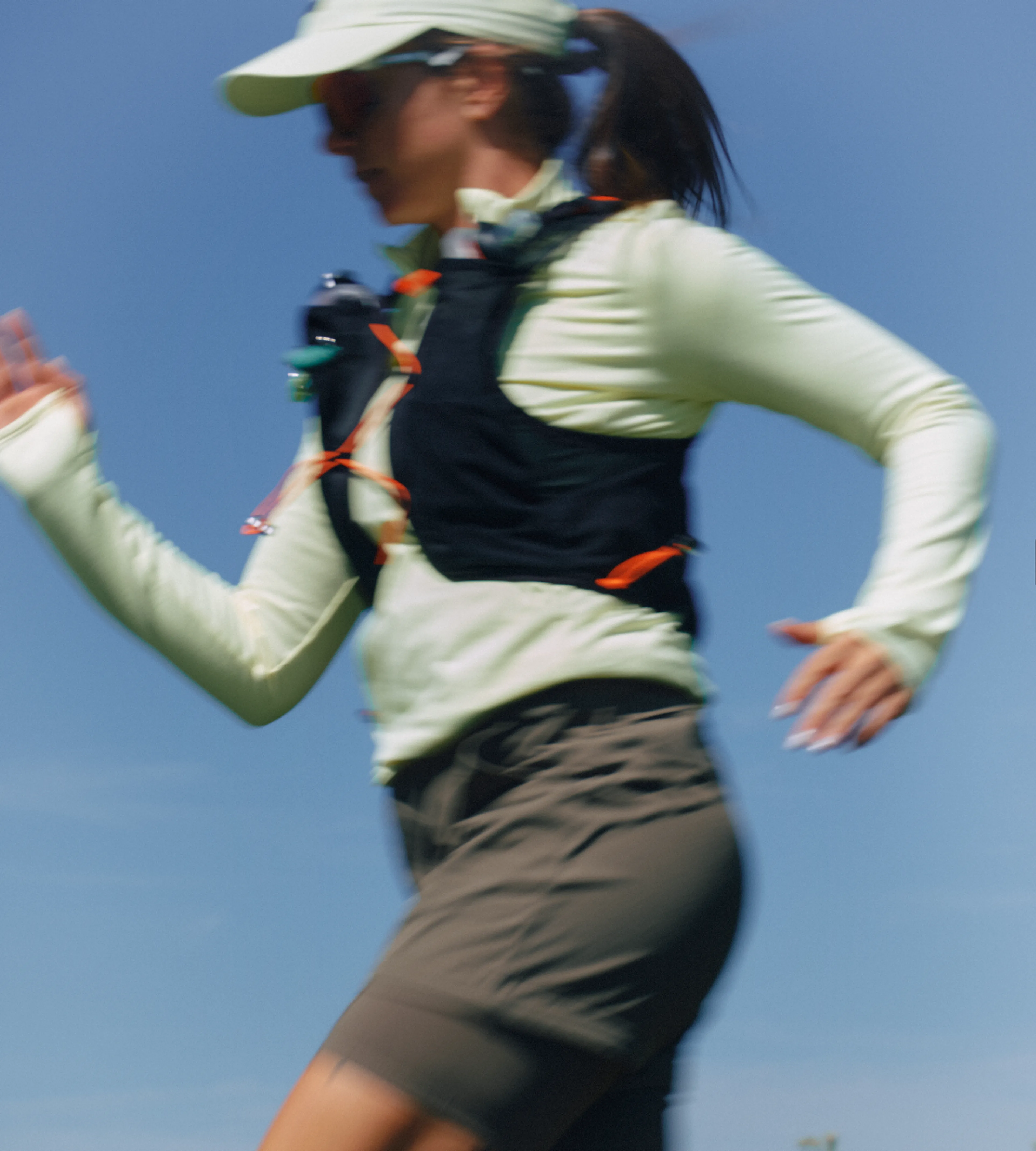 A woman in athletic gear runs outdoors on a sunny day, wearing a light cap, sunglasses, long-sleeve shirt, vest, and shorts. The image is slightly blurred, emphasizing motion against a clear blue sky.
