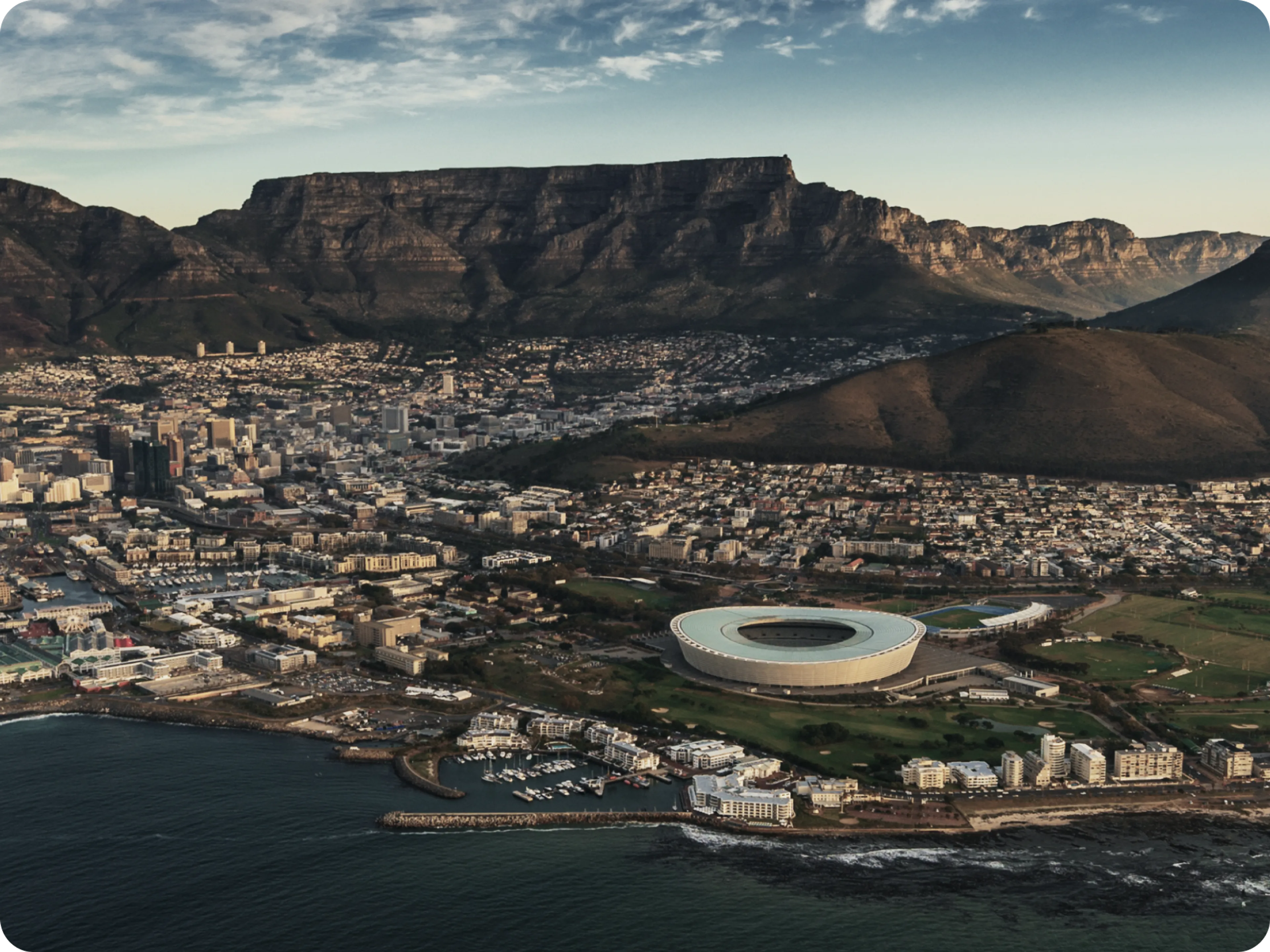 Aerial view of Cape Town, South Africa, showing the city, Cape Town Stadium, the coastline, and Table Mountain in the background under a partly cloudy sky.