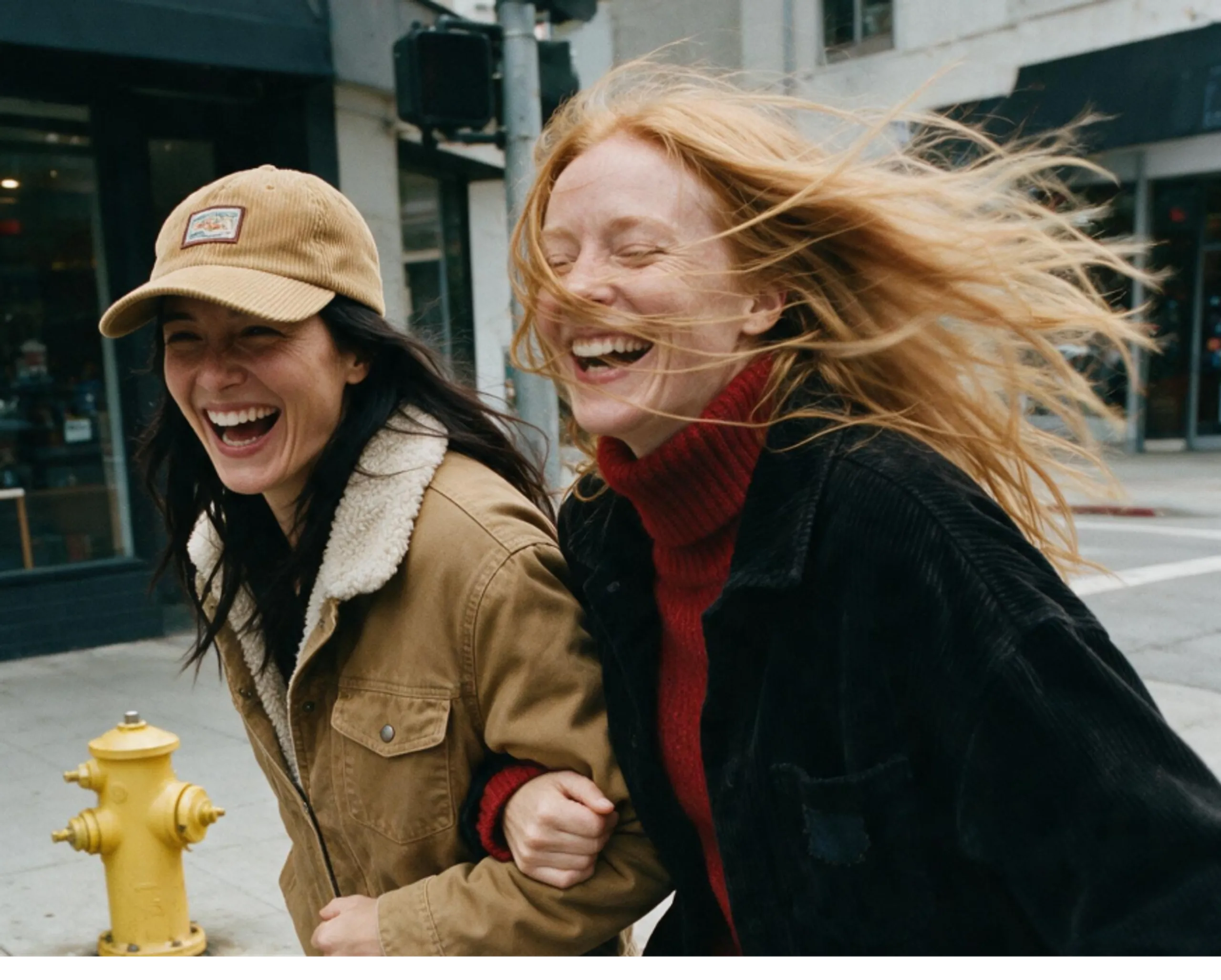 Two women outdoors laughing together, one wearing a tan jacket and cap, the other with long red hair in a black jacket. They hold arms while walking past a yellow fire hydrant on a city street.