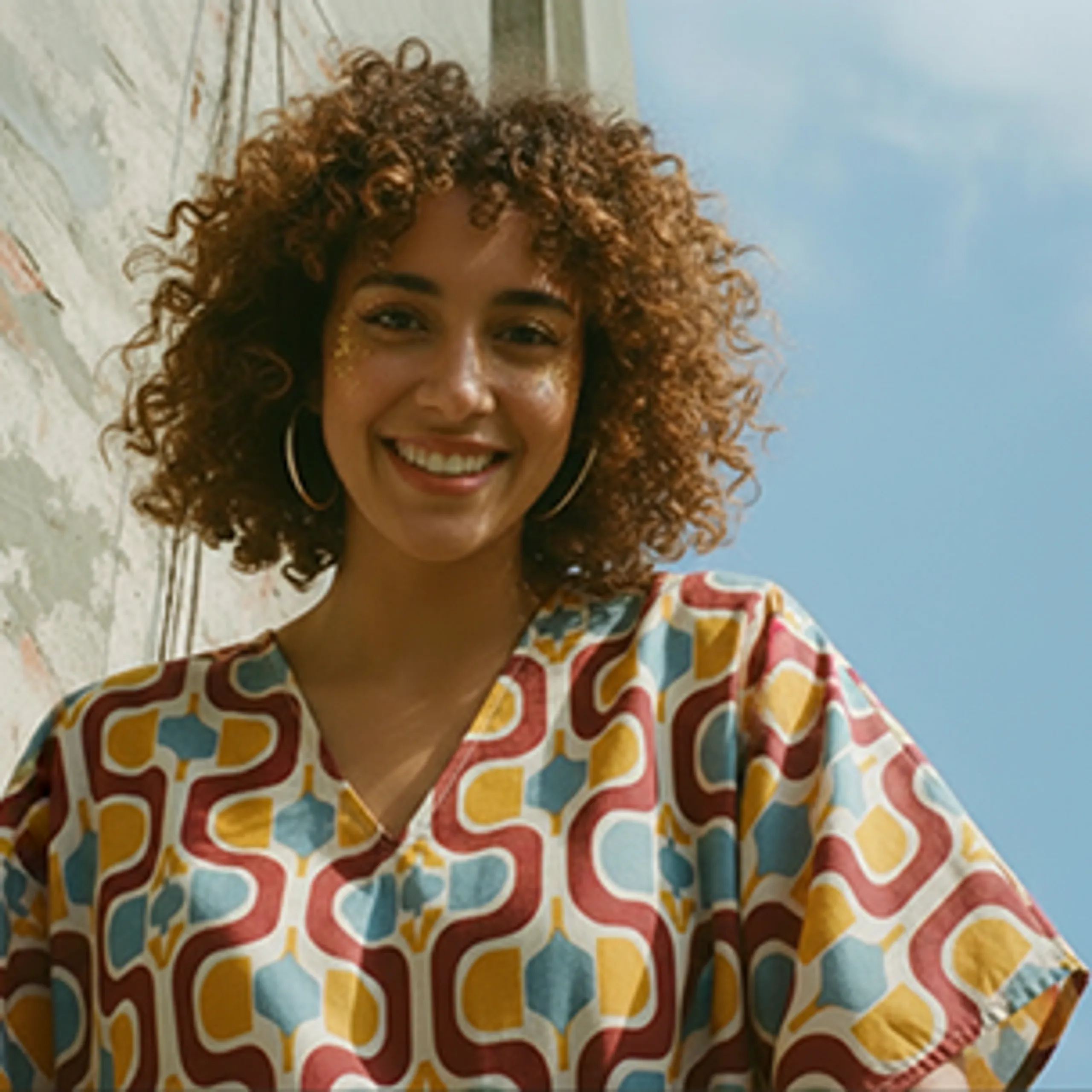 A young woman with curly hair, wearing large hoop earrings and a colorful geometric-patterned shirt, smiles outdoors near a weathered wall under a blue sky.