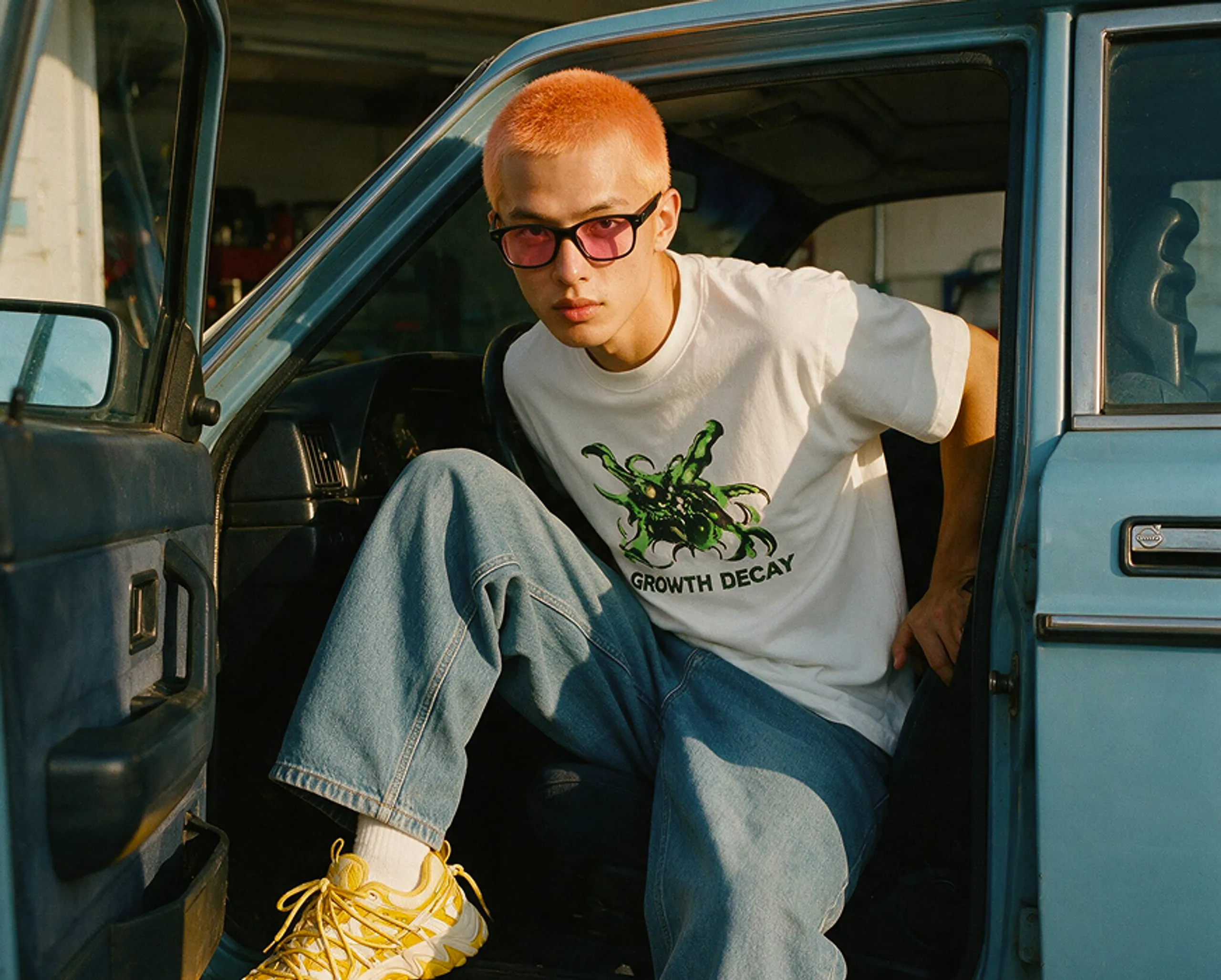 A person with short orange hair, pink sunglasses, and a white graphic T-shirt sits in the open door of a blue car, looking at the camera. They wear loose jeans and yellow sneakers.