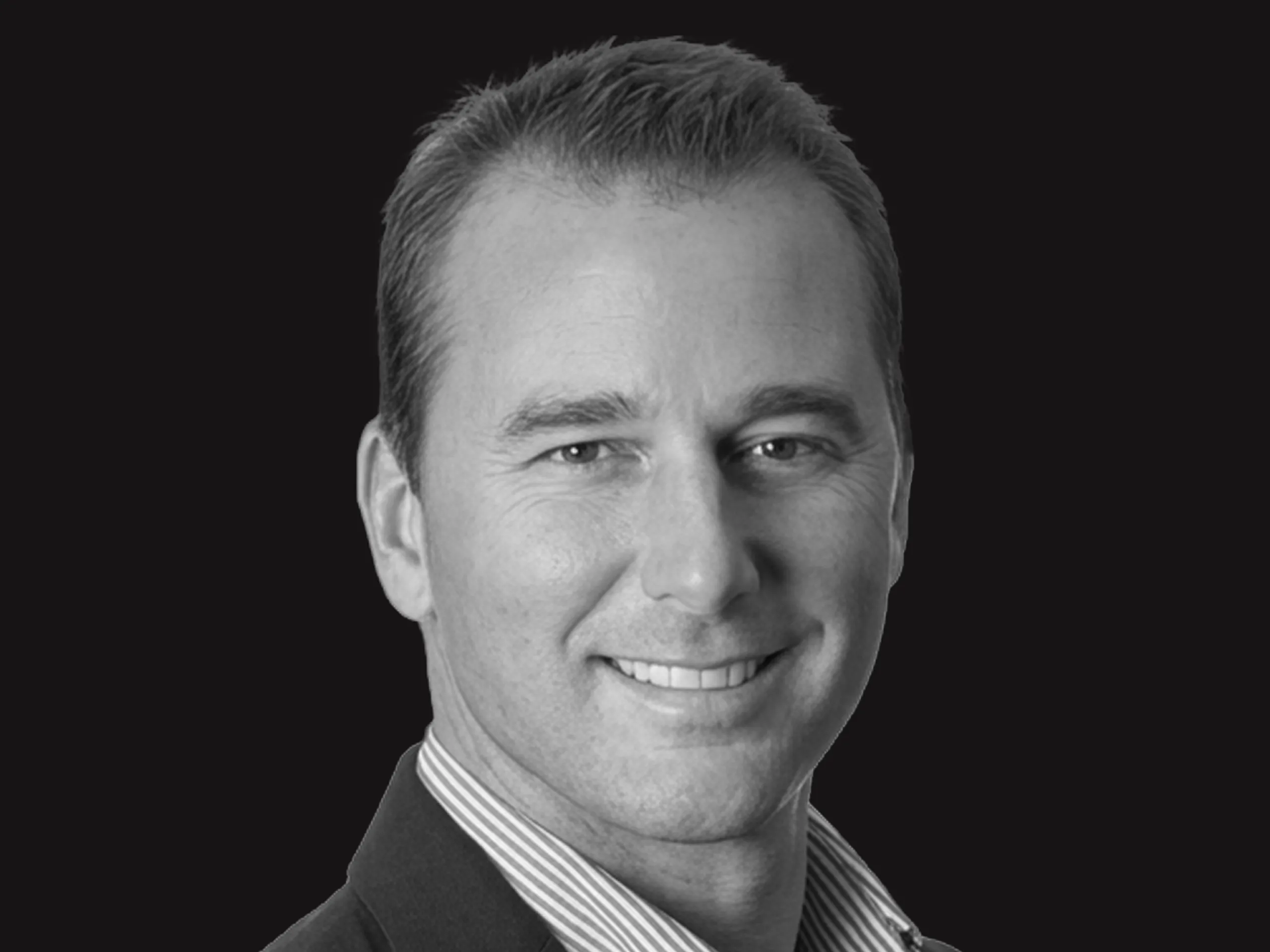A man with short hair smiles, wearing a collared shirt and jacket, posed against a plain black background in a black-and-white portrait.