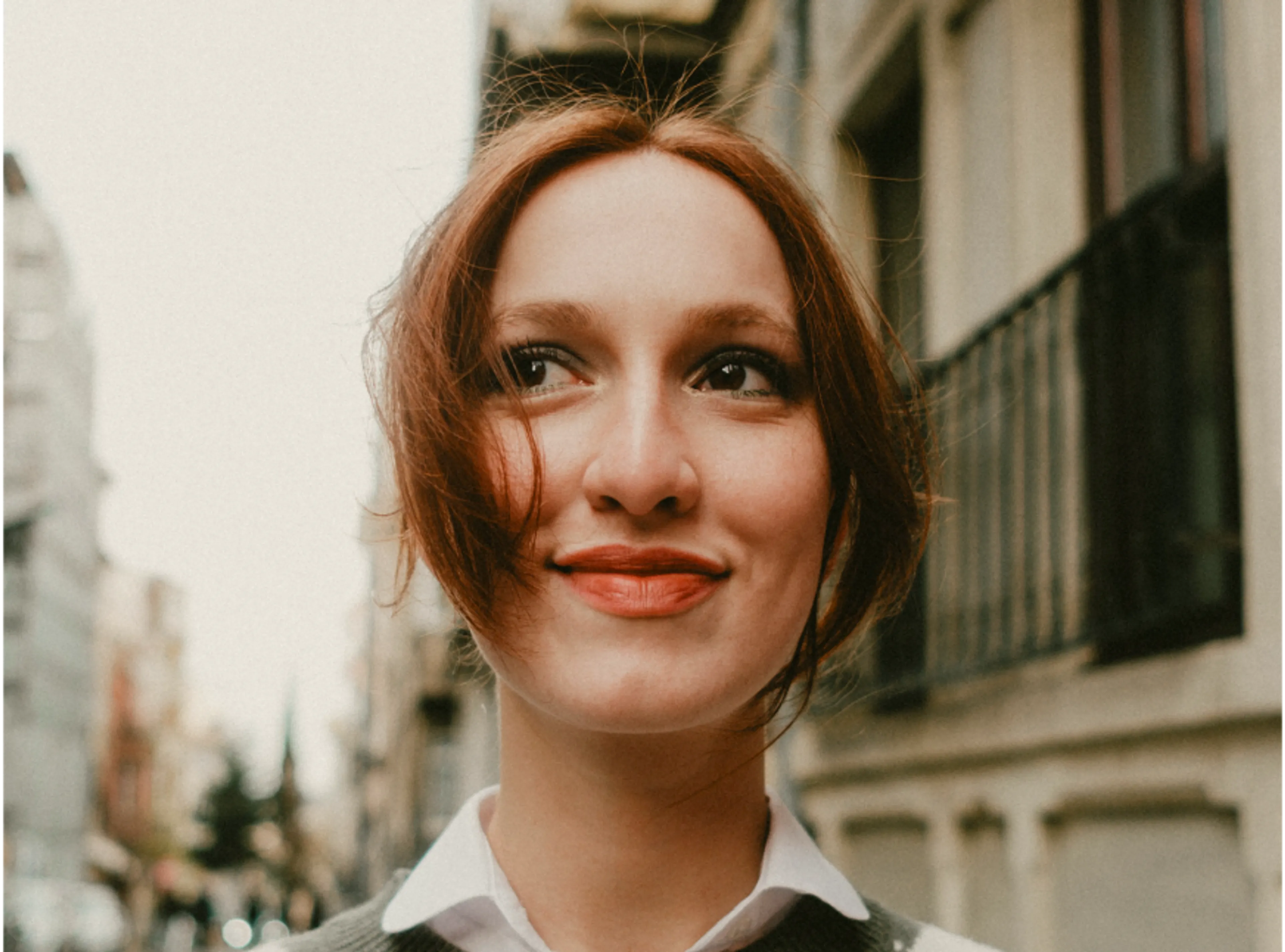 A young woman with reddish-brown hair and a white collared shirt smiles while standing outdoors in a city, with buildings and a tree-lined street in the background.