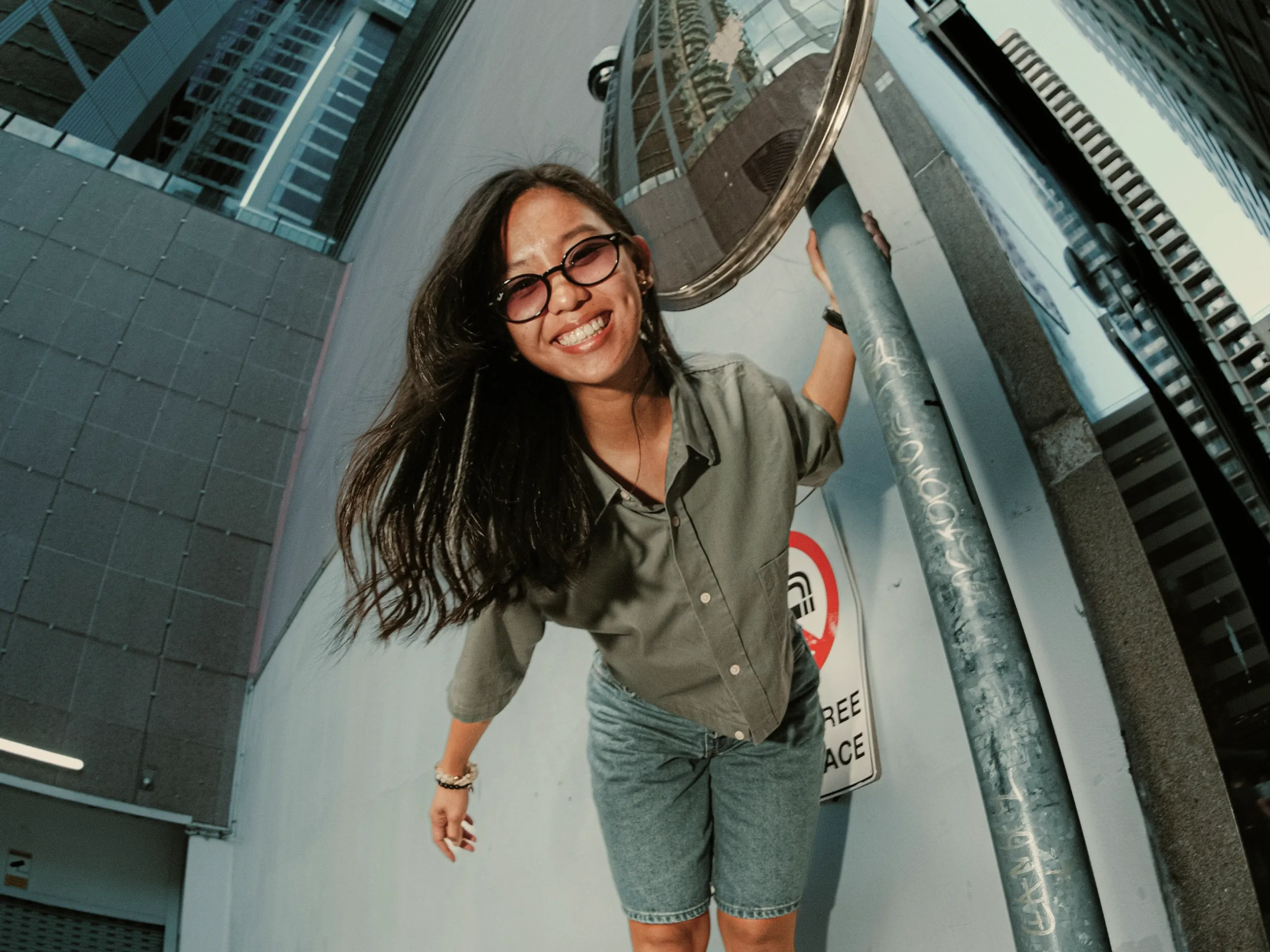 A smiling woman with glasses stands outdoors, leaning on a metal pole beside a convex mirror. She wears a green shirt and denim shorts, with buildings reflected in the mirror behind her.