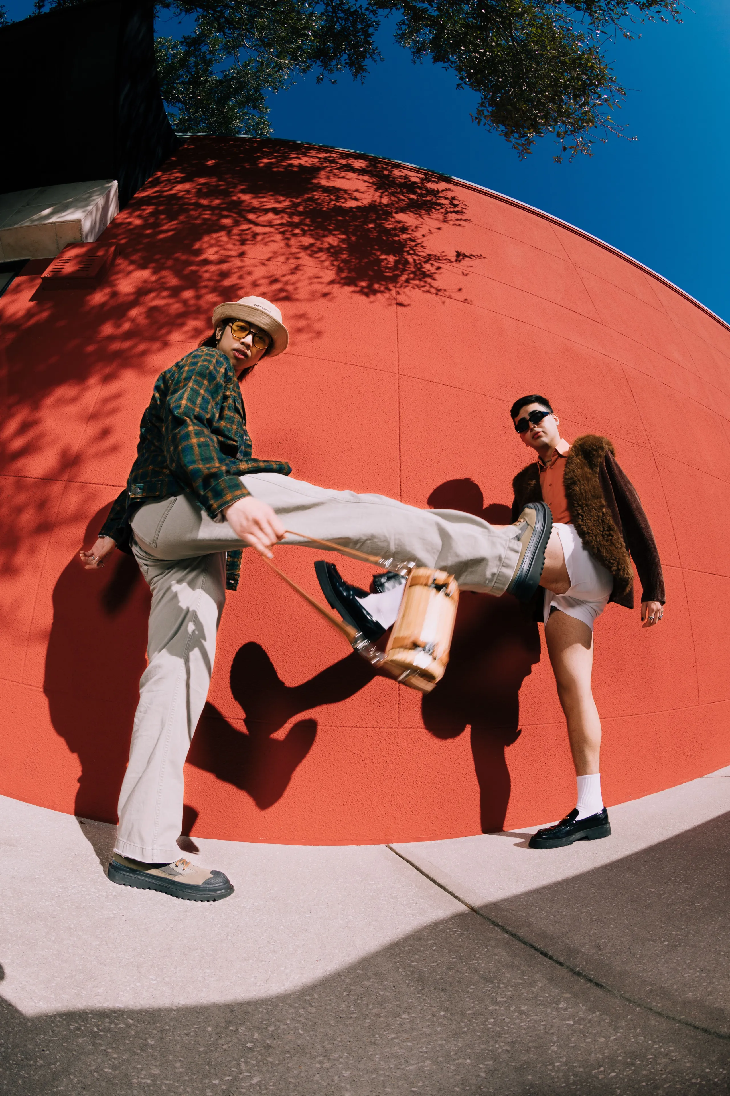 Two stylish men pose outdoors against a bright red wall. One man in light pants and a hat lifts his leg toward the camera, while the other, in shorts and sunglasses, stands confidently beside him.