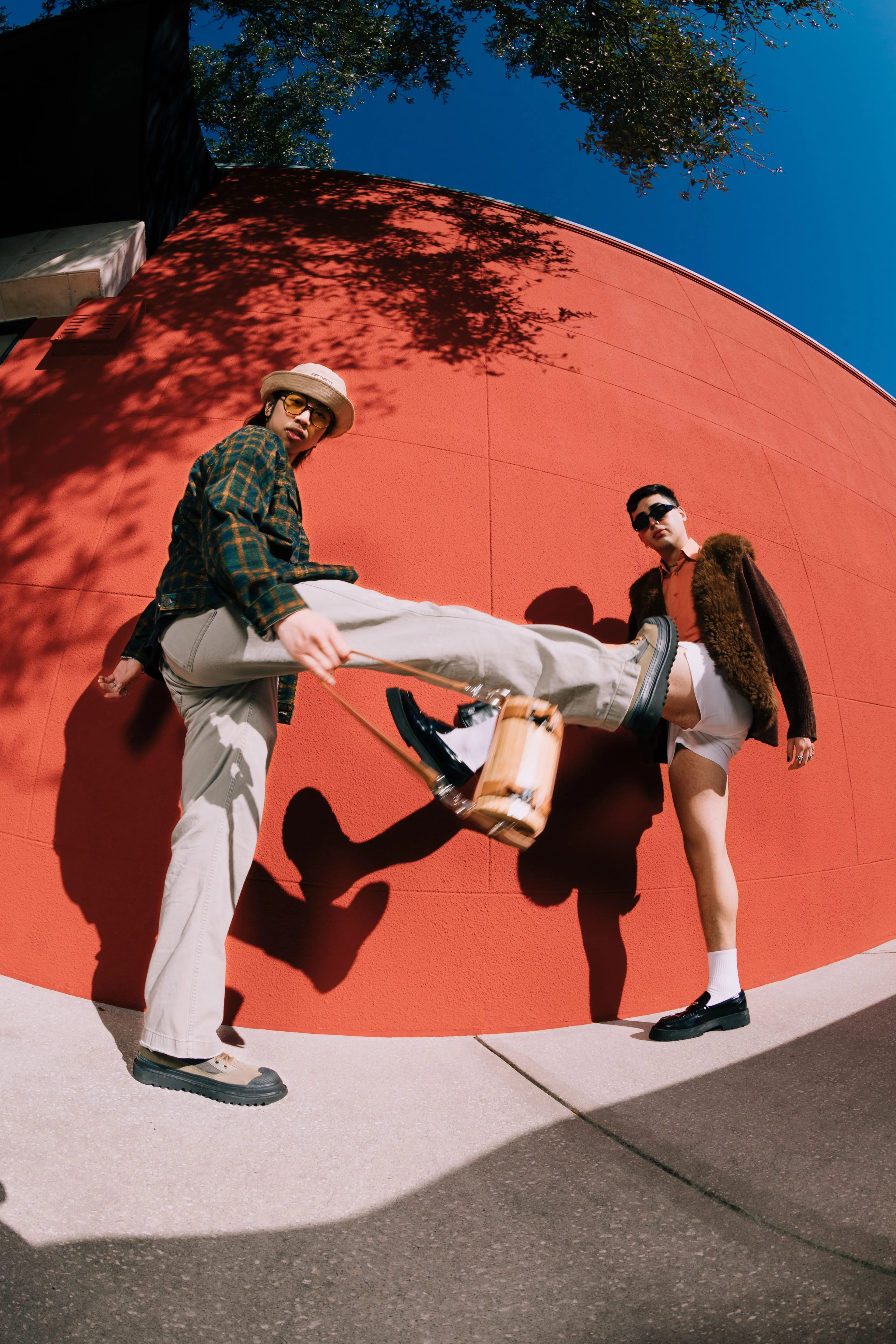 Two stylish men pose outdoors against a bright red wall. One man in light pants and a hat lifts his leg toward the camera, while the other, in shorts and sunglasses, stands confidently beside him.