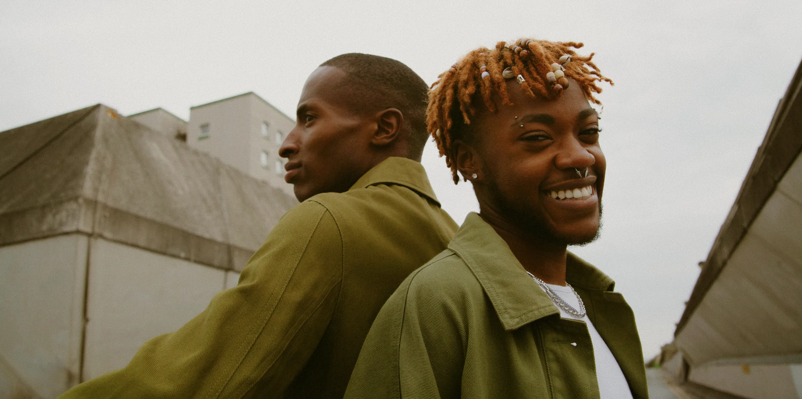 Two young men stand back to back, both wearing green jackets. One faces sideways looking ahead, while the other smiles at the camera. Urban buildings and a cloudy sky are in the background.