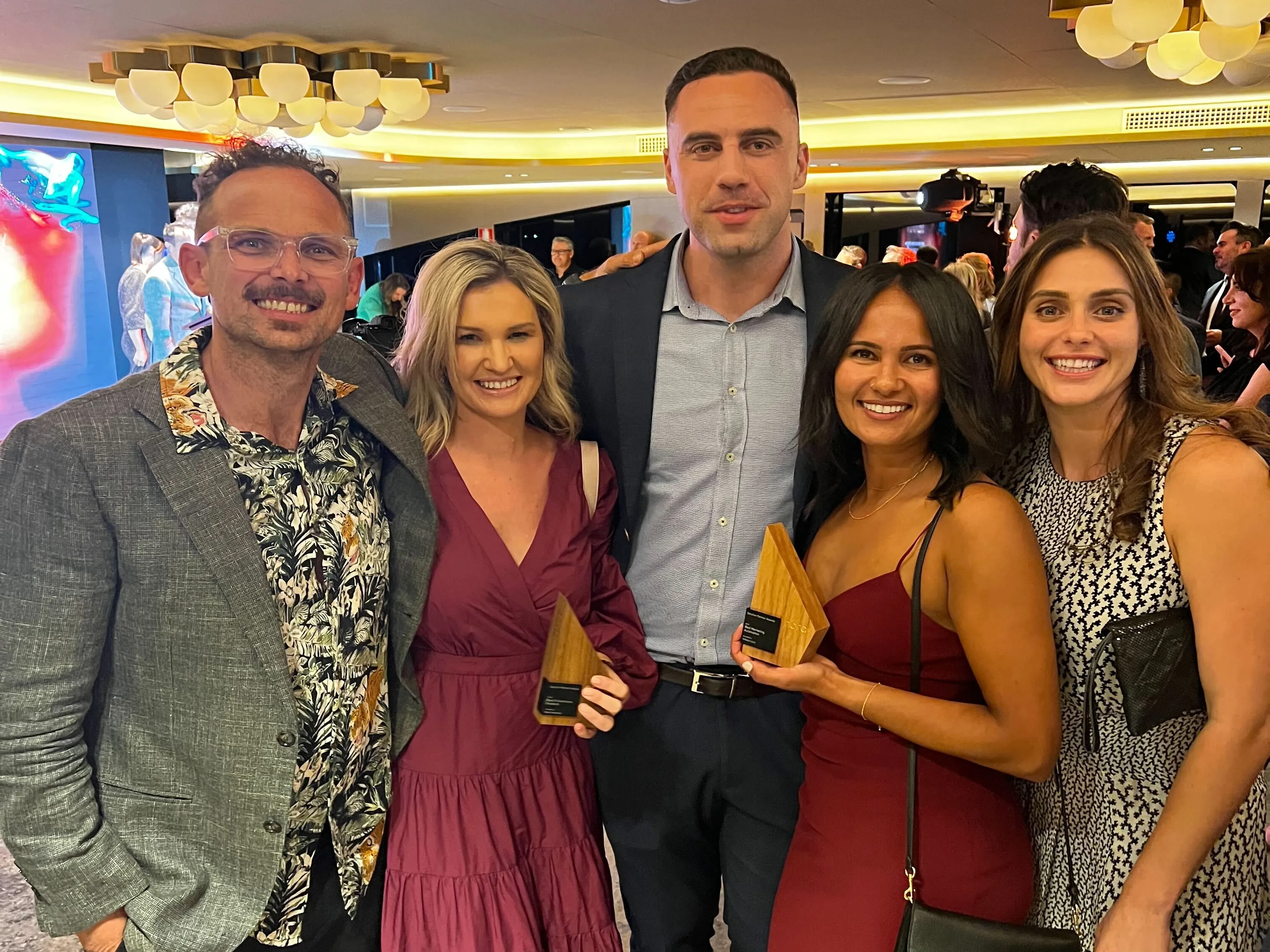Five people dressed formally smile at an indoor event. Two women in the front hold small wooden awards. The background shows other attendees and warm lighting.