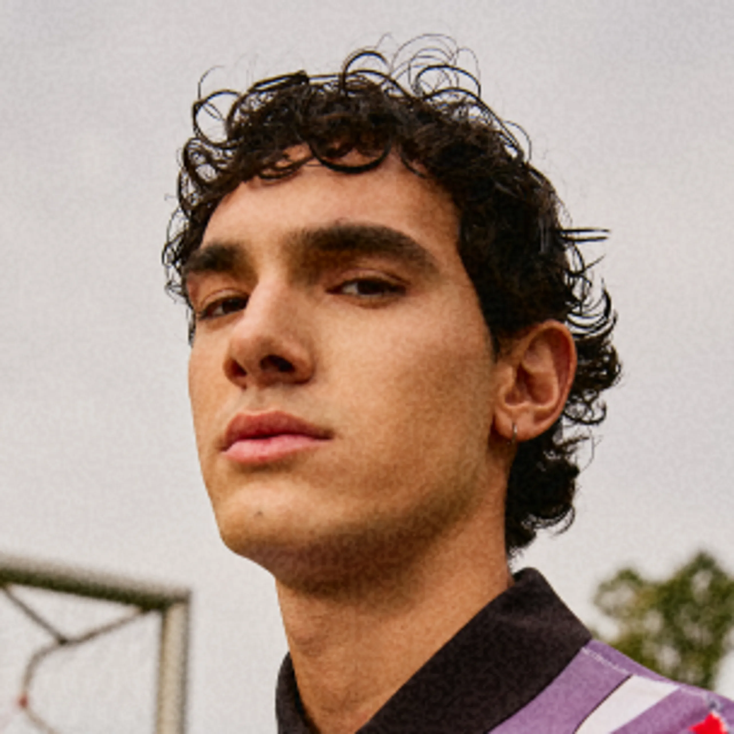 A young person with short, curly dark hair and a small hoop earring looks confidently at the camera outdoors, with a blurred background featuring part of a basketball hoop.