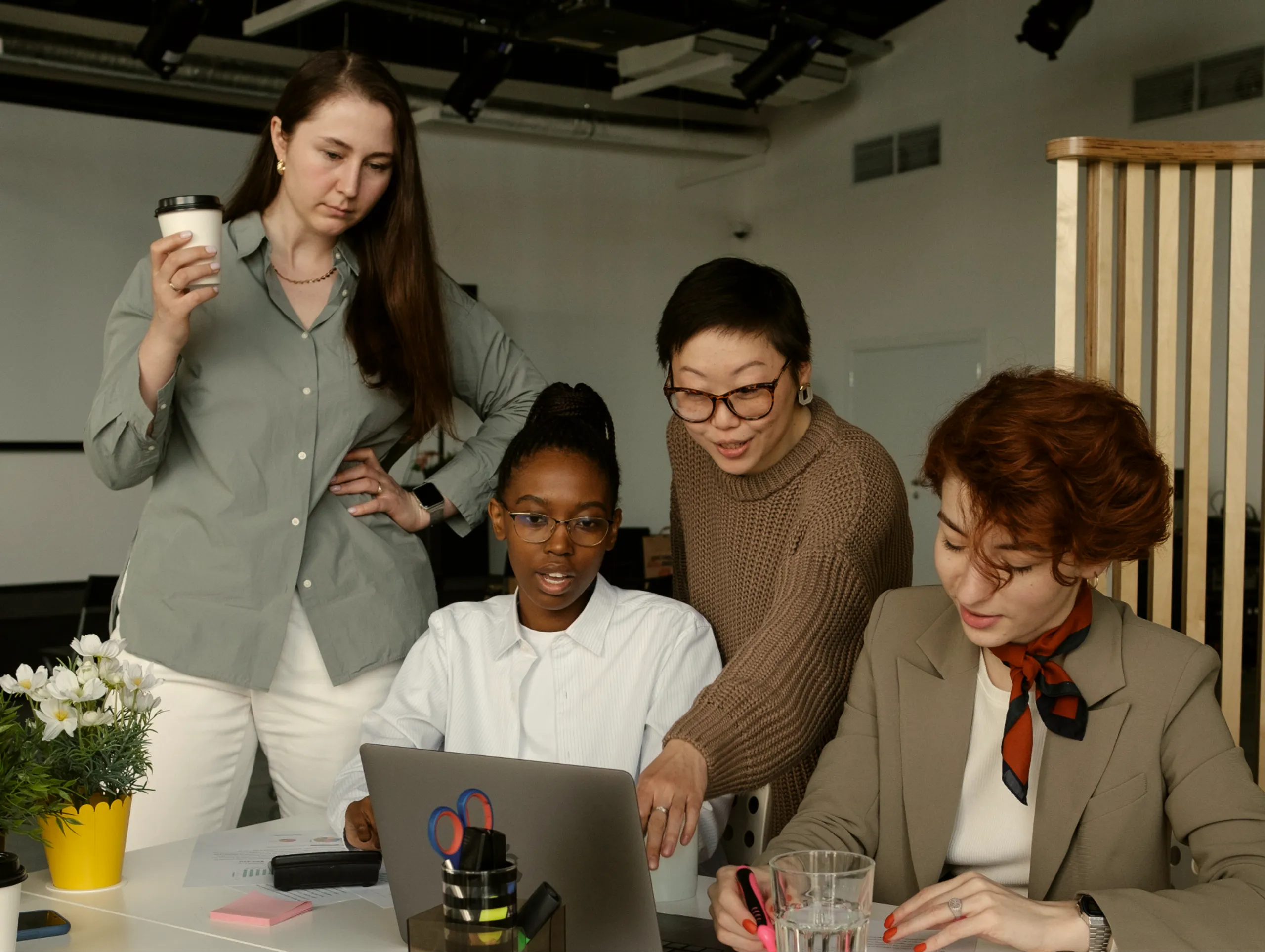 Four women collaborate at a desk with a laptop. One holds a coffee cup, another points at the screen, while the others watch and take notes. Office supplies and a small plant are on the table.