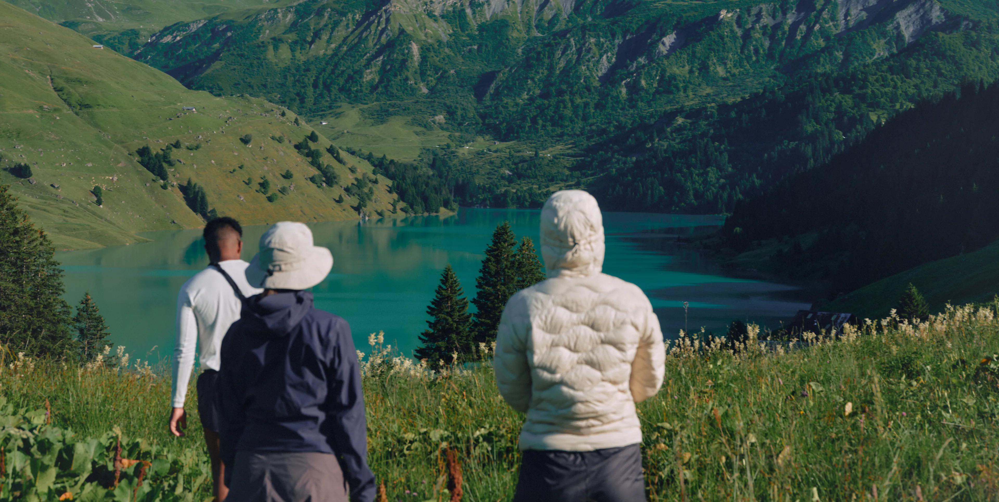 Three people in outdoor clothing face a turquoise lake surrounded by green mountains and forests, standing in a grassy field under clear daylight.