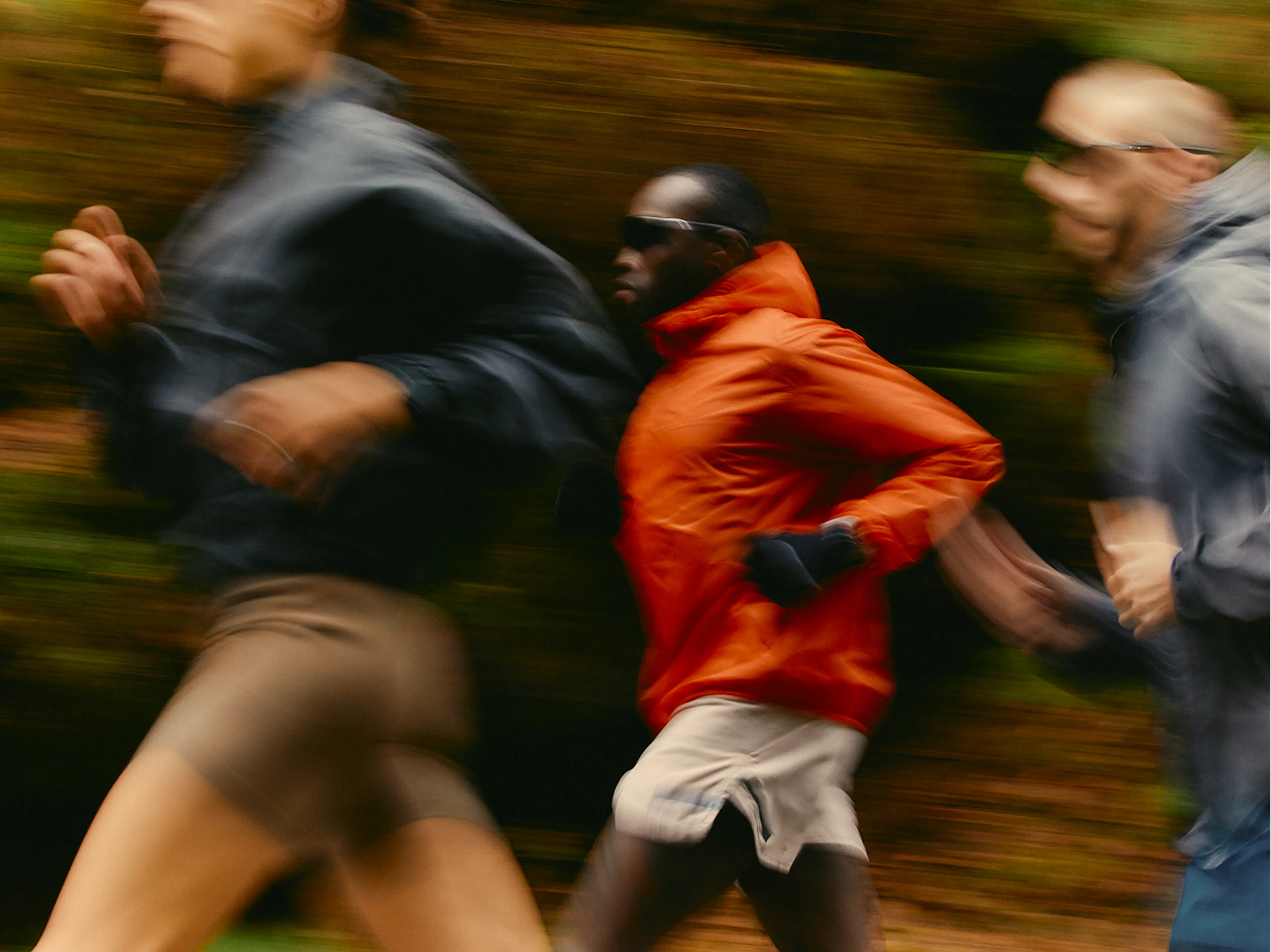 Three people are running outdoors. The image is slightly blurred to show motion. The central runner wears a red jacket and sunglasses, while the others wear darker clothing. The background appears as a blur of green and brown.