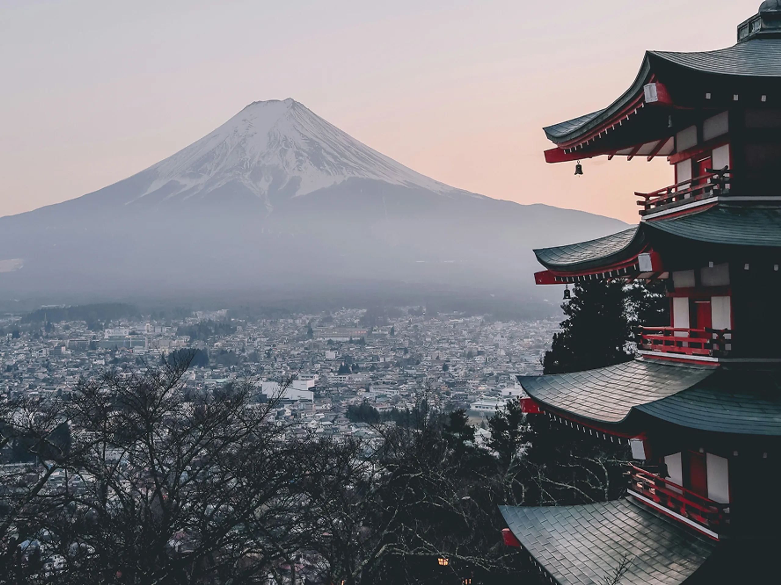 A five-story pagoda in the foreground with Mount Fuji in the background at sunset, overlooking a city partially covered in mist and surrounded by bare trees.