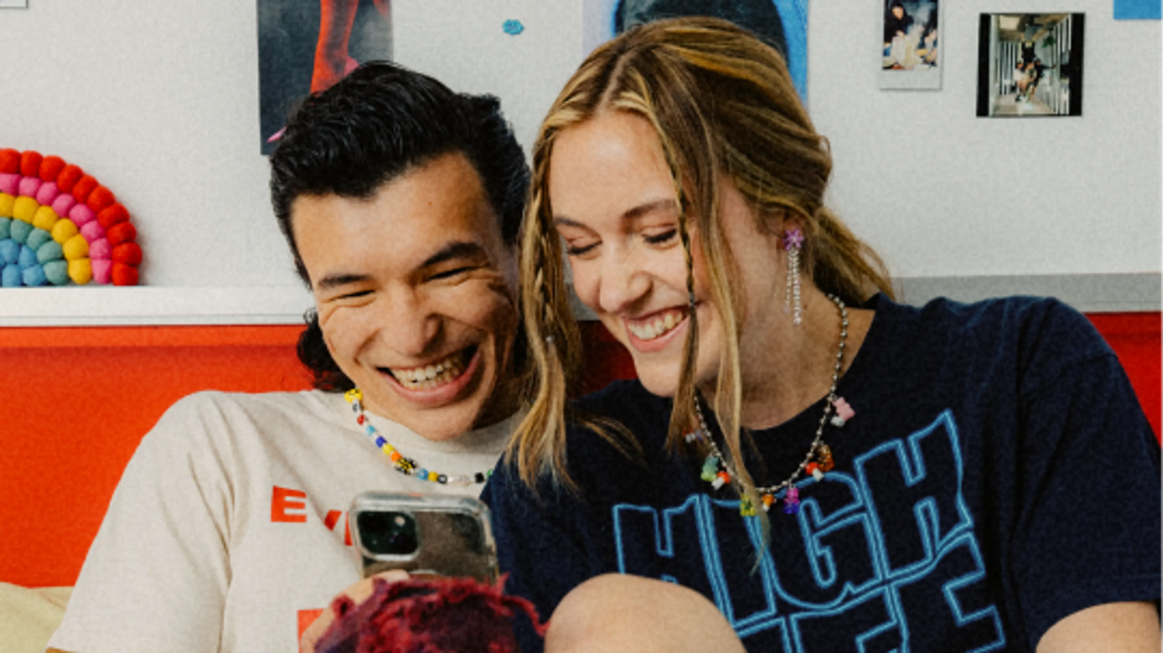 Two people sitting close together on a bed, smiling and looking at a smartphone. The background includes colorful decorations and photos on the wall.