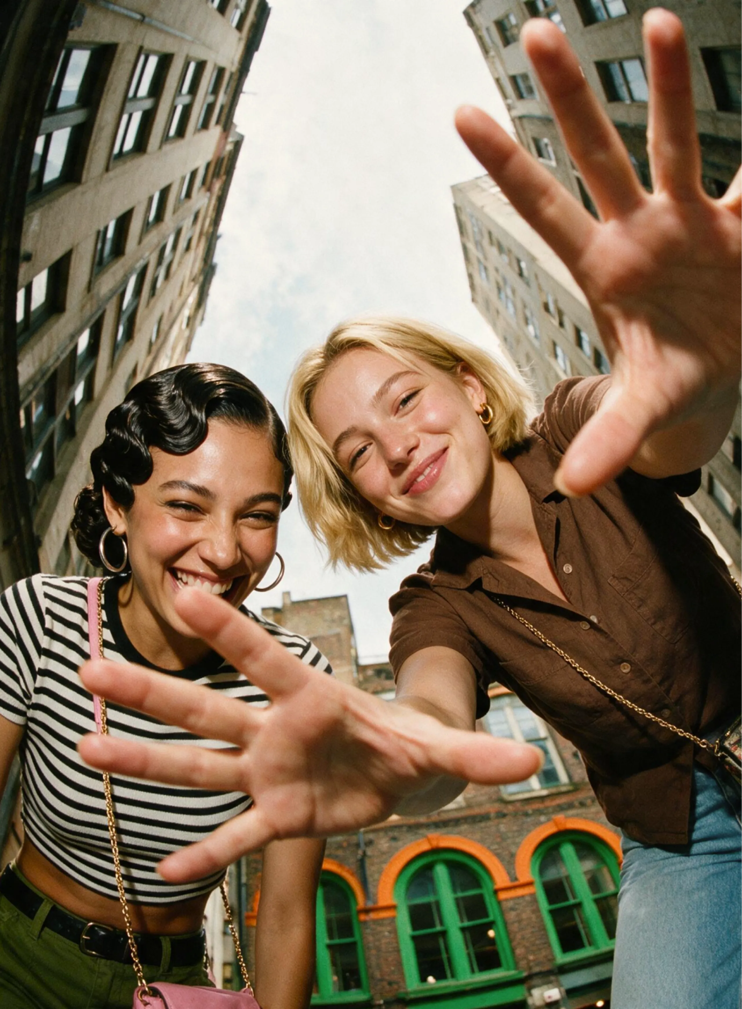 Two young women smile and reach toward the camera while standing between tall city buildings on a sunny day. The photo is taken with a fisheye lens, creating a playful, distorted perspective.