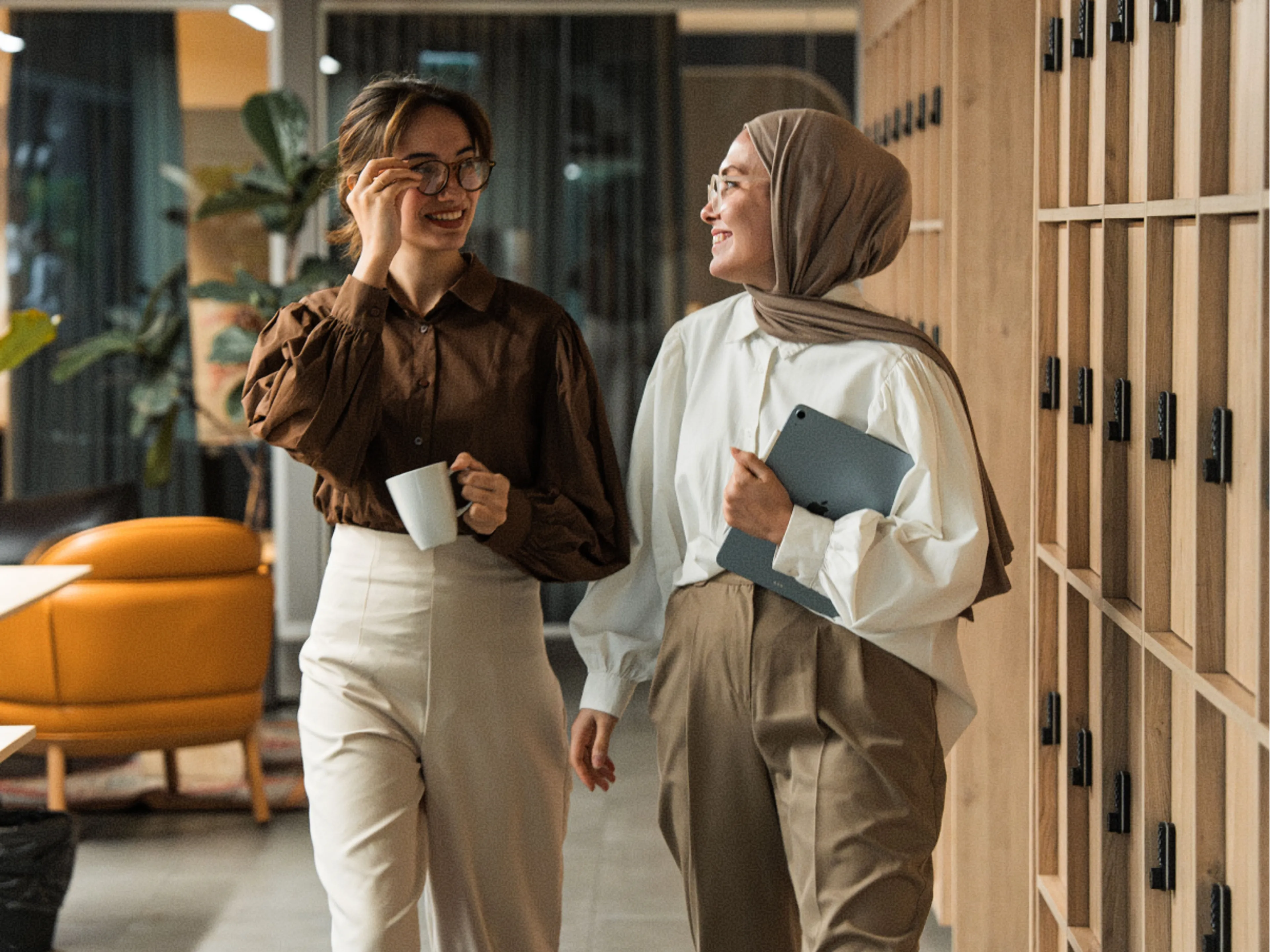Two women walking and smiling in an office hallway; one holds a coffee mug and wears glasses, while the other, wearing a hijab, holds a tablet. They appear to be engaged in a friendly conversation.