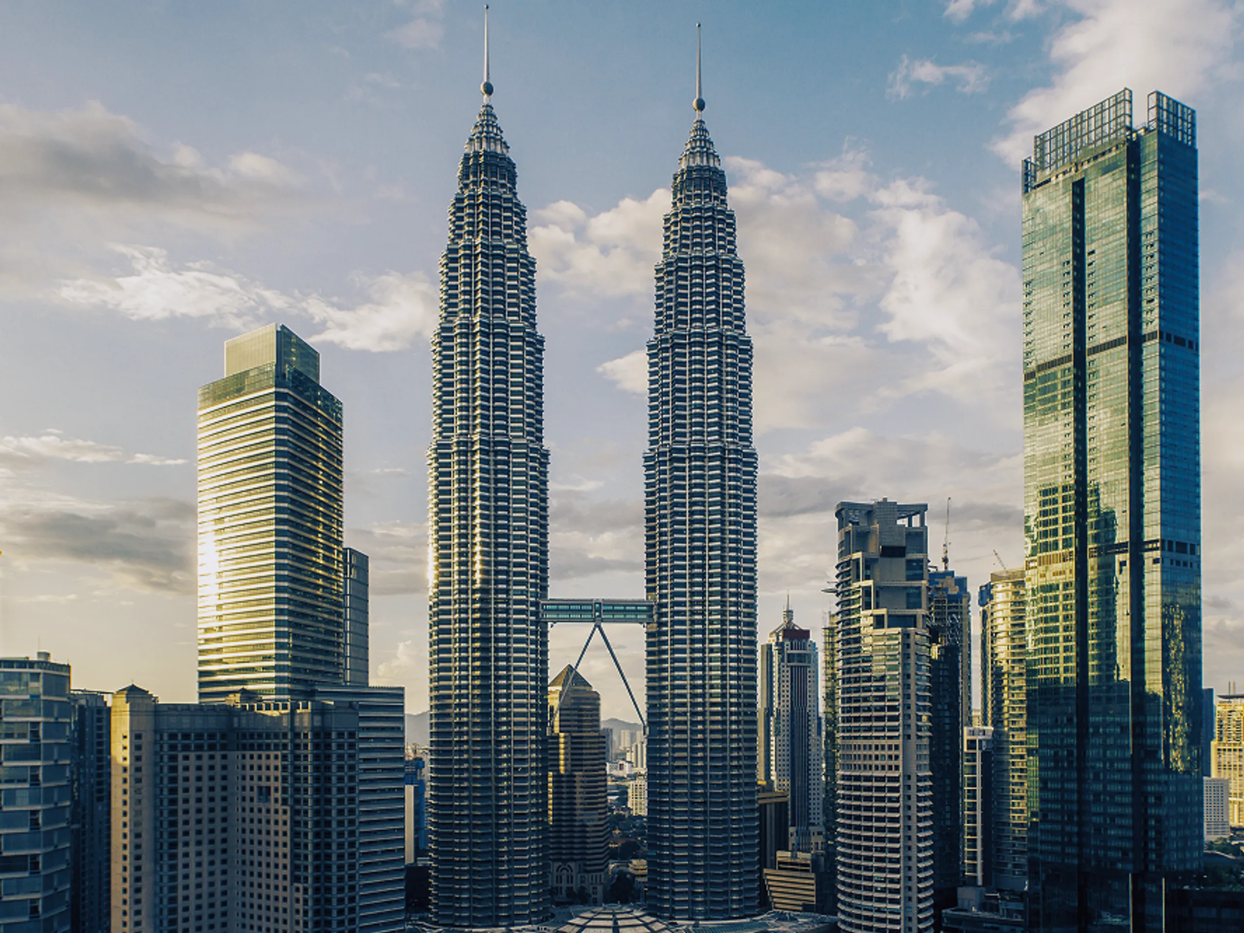 A city skyline at sunset features the twin Petronas Towers and modern skyscrapers, with a partly cloudy sky in the background.