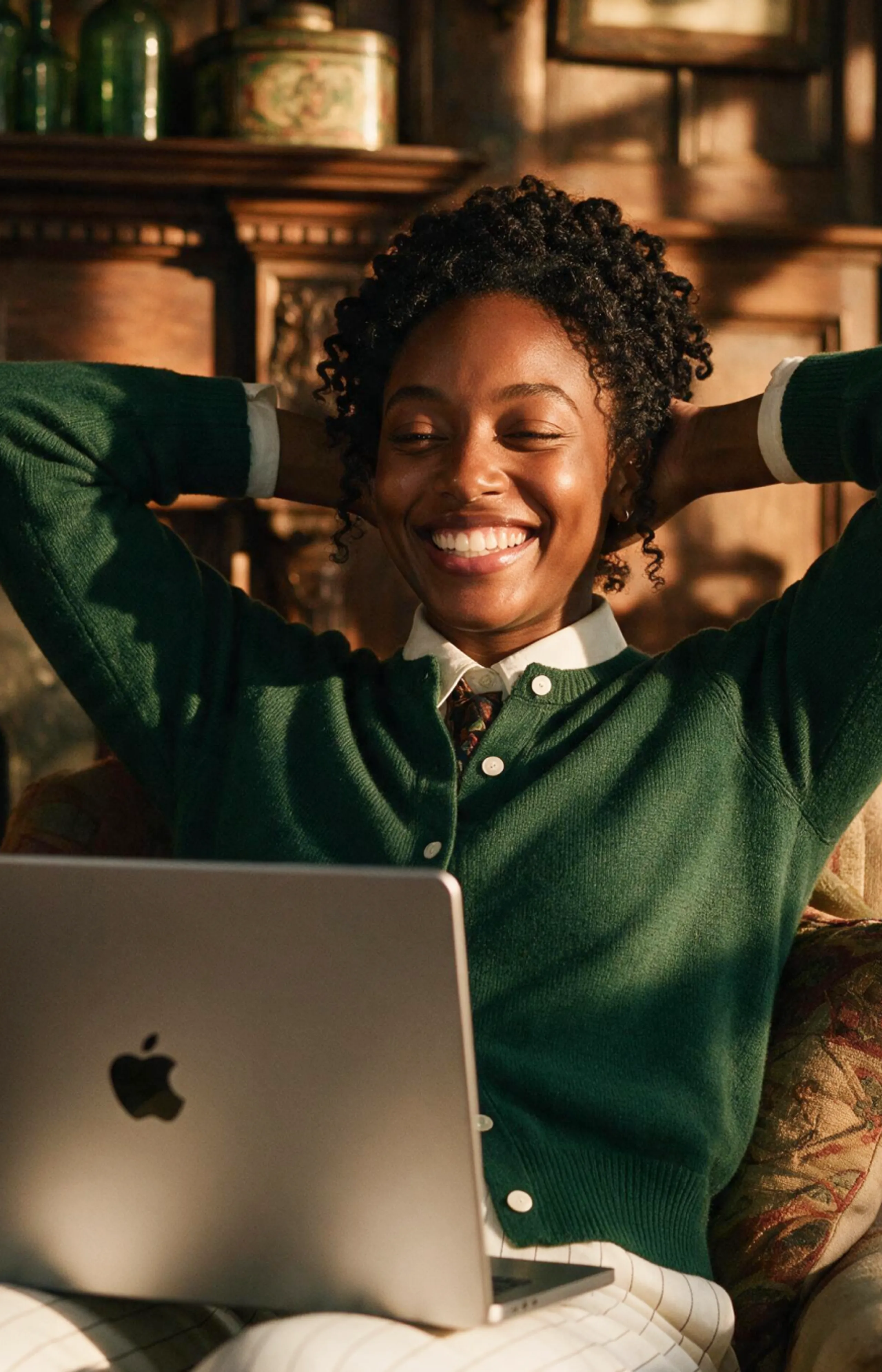A smiling person in a green sweater relaxes with hands behind their head while looking at a laptop. Warm sunlight streams in, creating a cozy atmosphere.