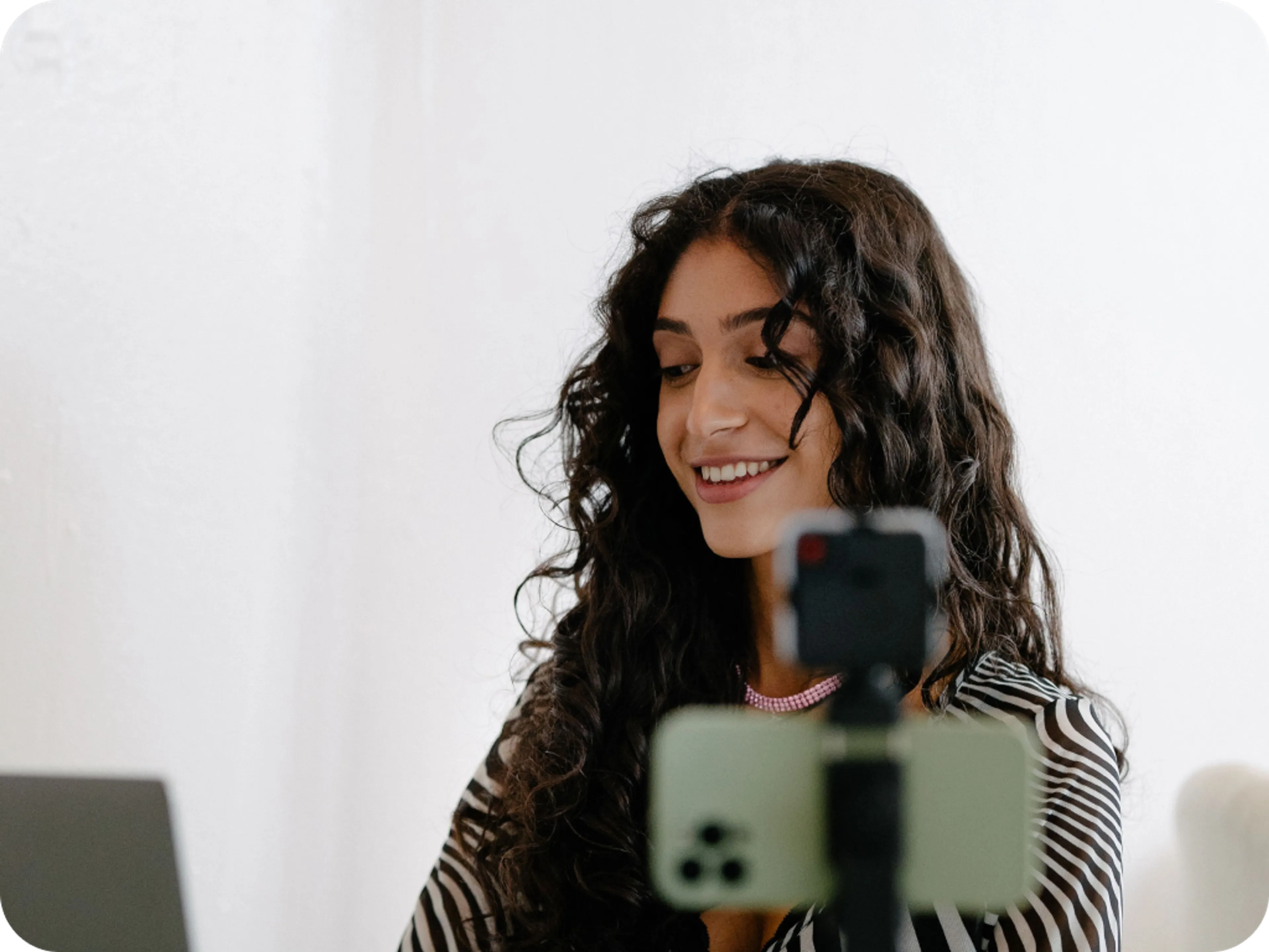 A young woman with long curly hair smiles while recording a video on her smartphone mounted to a tripod.