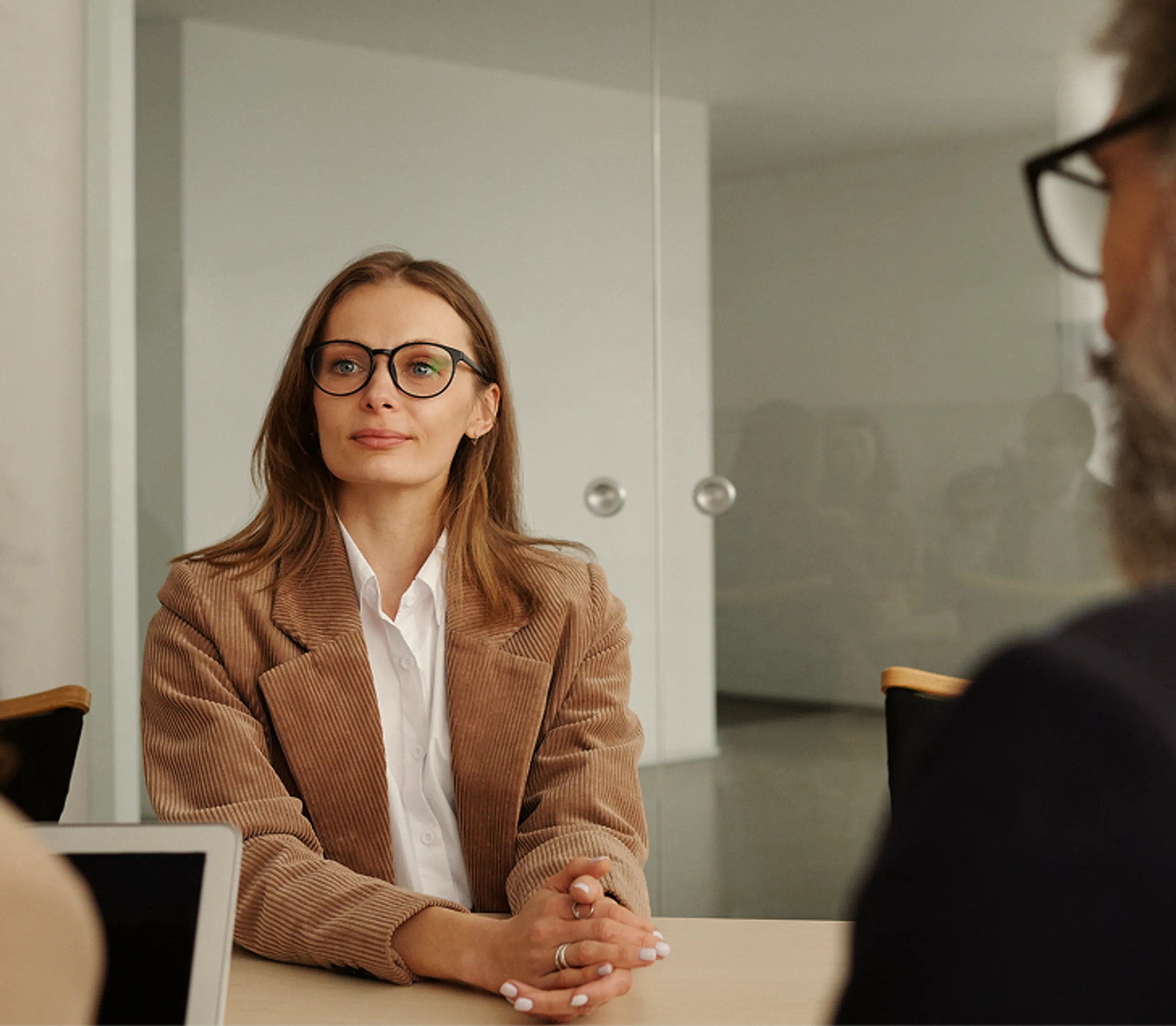 A woman with glasses and a brown blazer sits at a table during a meeting or interview, facing two blurred people across from her in a modern office setting.
