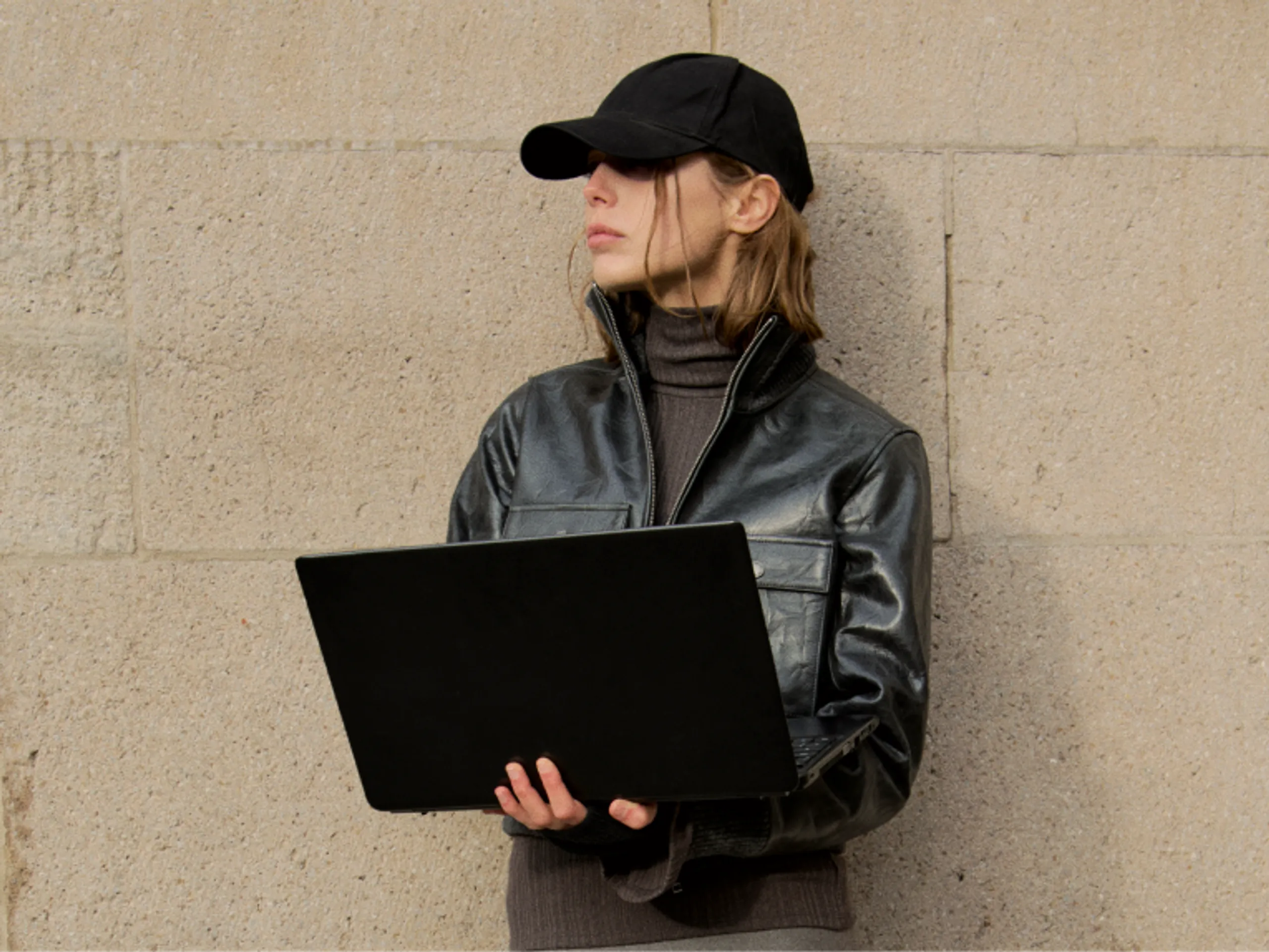 A person in a black cap and leather jacket holds a laptop while standing against a beige stone wall, looking to the side.