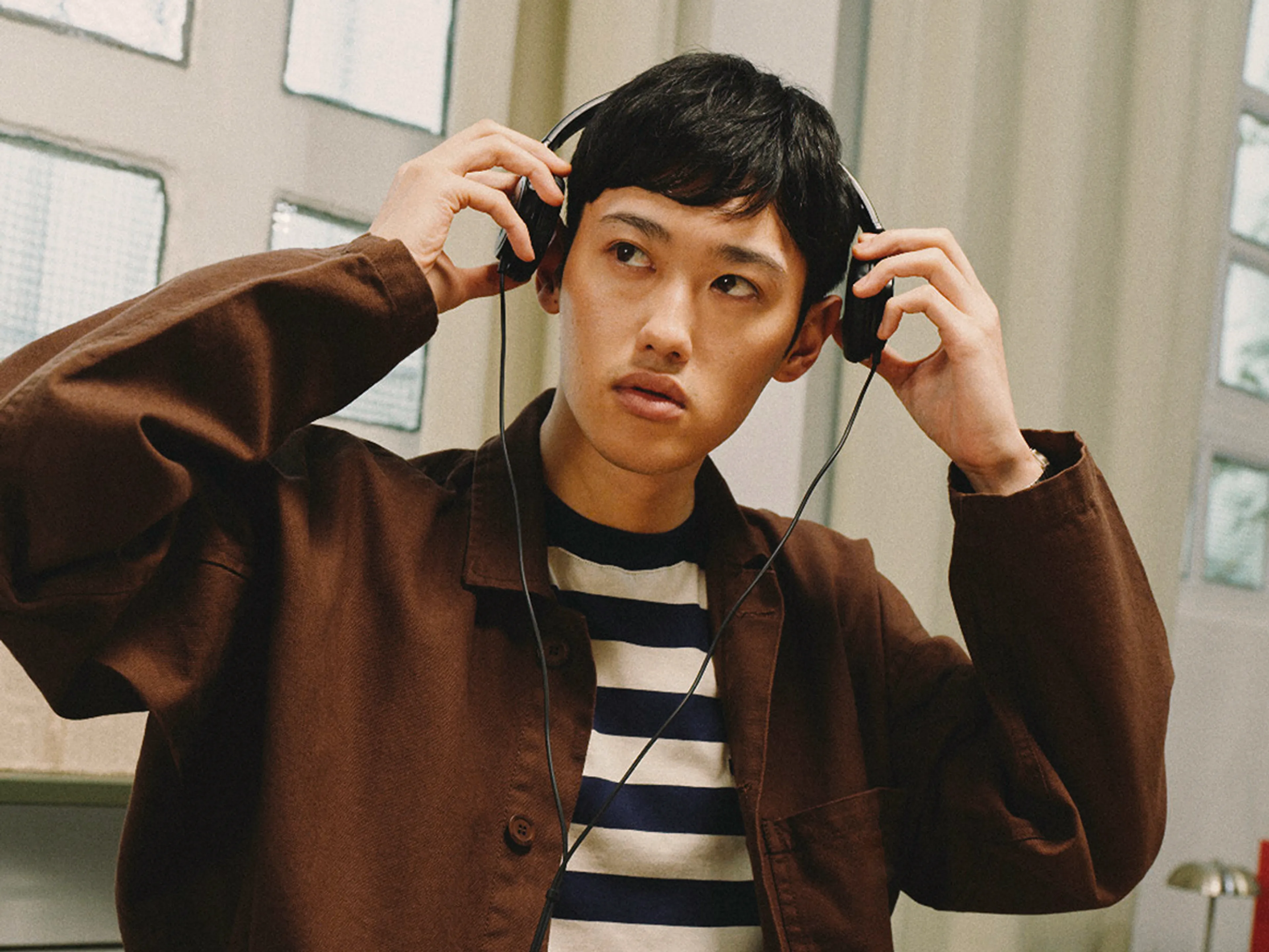 A person wearing a brown jacket and striped shirt adjusts black headphones on their head, looking to the side. They are indoors with windows and light curtains in the background.