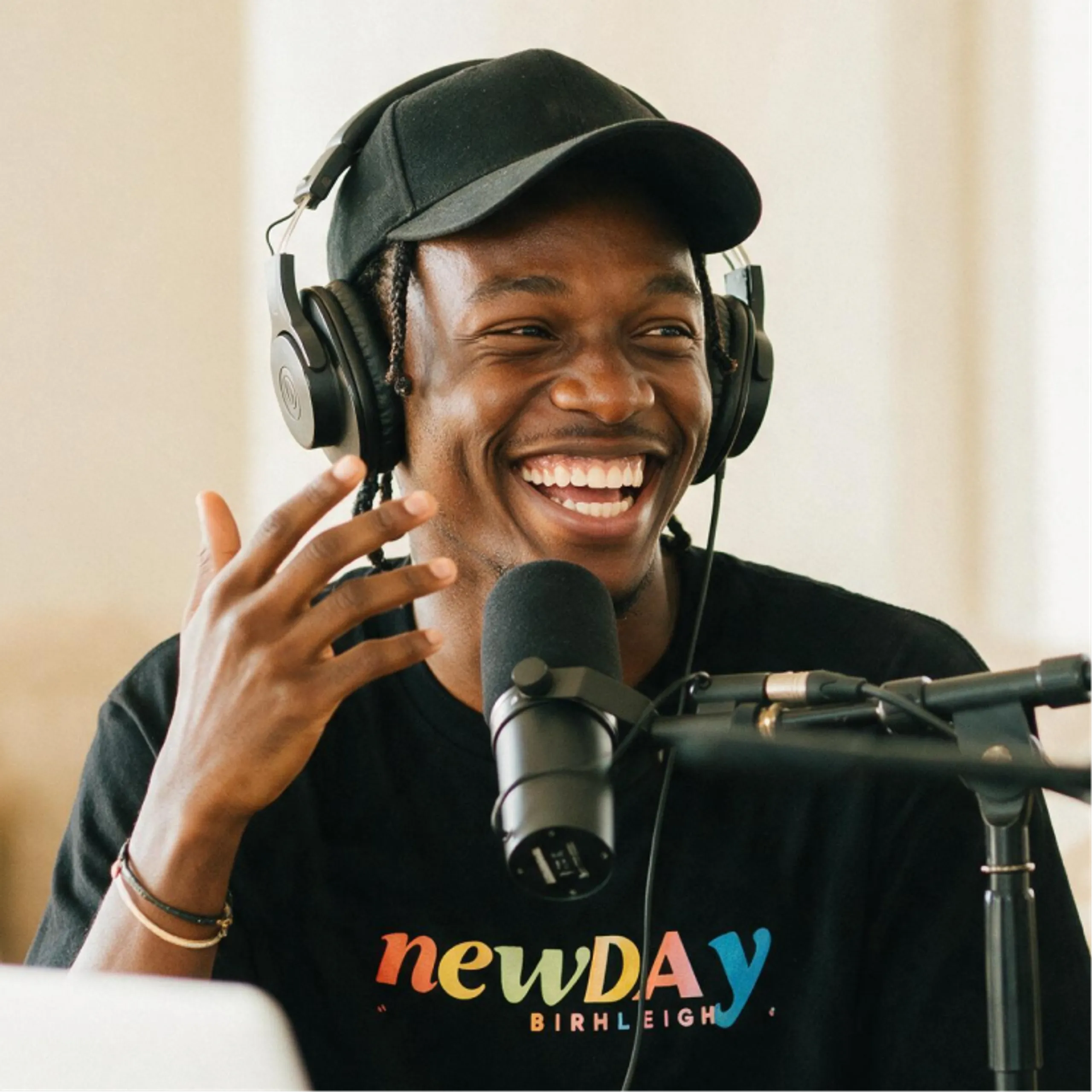 A smiling person wearing headphones and a black cap speaks into a microphone. They are wearing a black newDAY T-shirt and sitting in front of audio equipment, likely recording a podcast or radio show.
