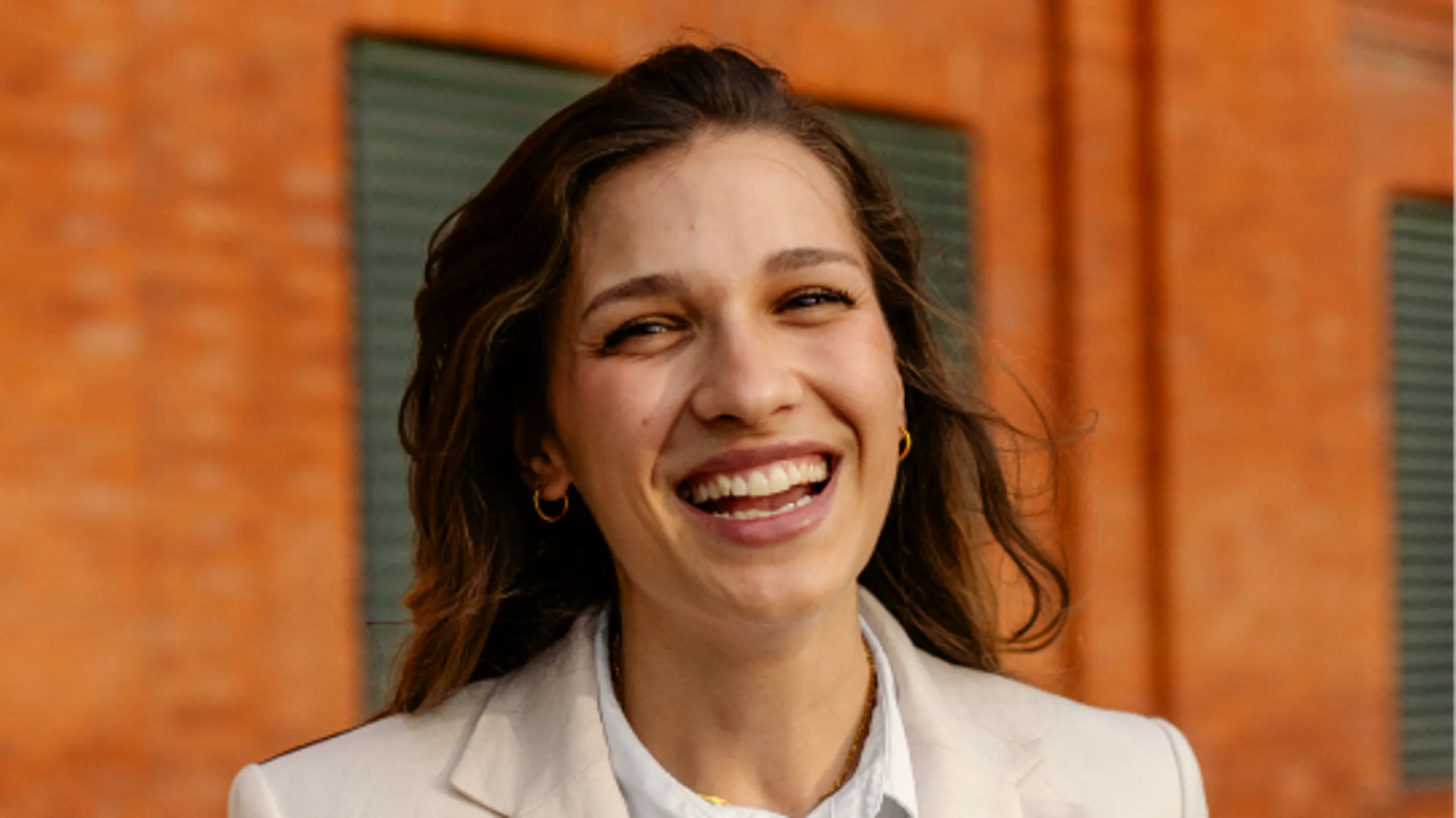 A woman with long brown hair smiles warmly at the camera, wearing a light-colored blazer, standing in front of an orange brick wall with green shutters.