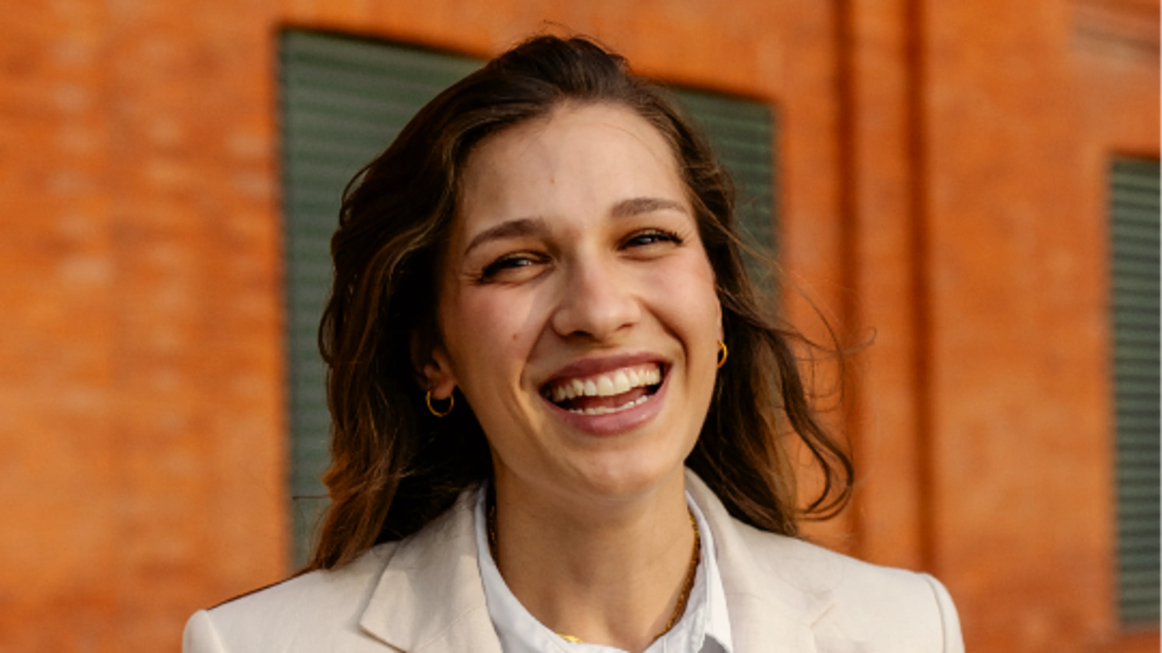 A woman with long brown hair smiles warmly at the camera, wearing a light-colored blazer, standing in front of an orange brick wall with green shutters.