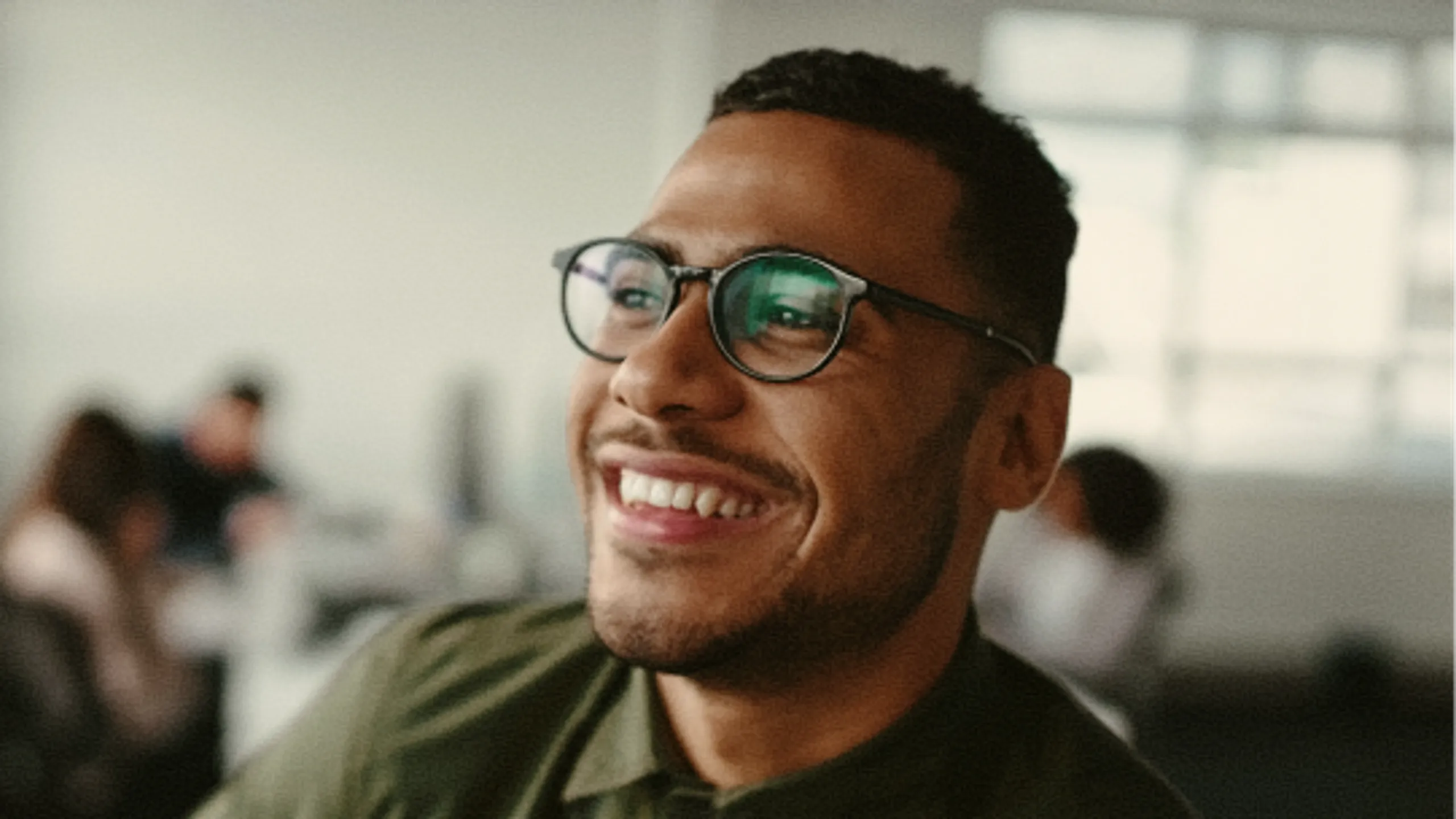 A smiling man with short hair and glasses looks off-camera in an office setting, with blurred people and windows in the background.