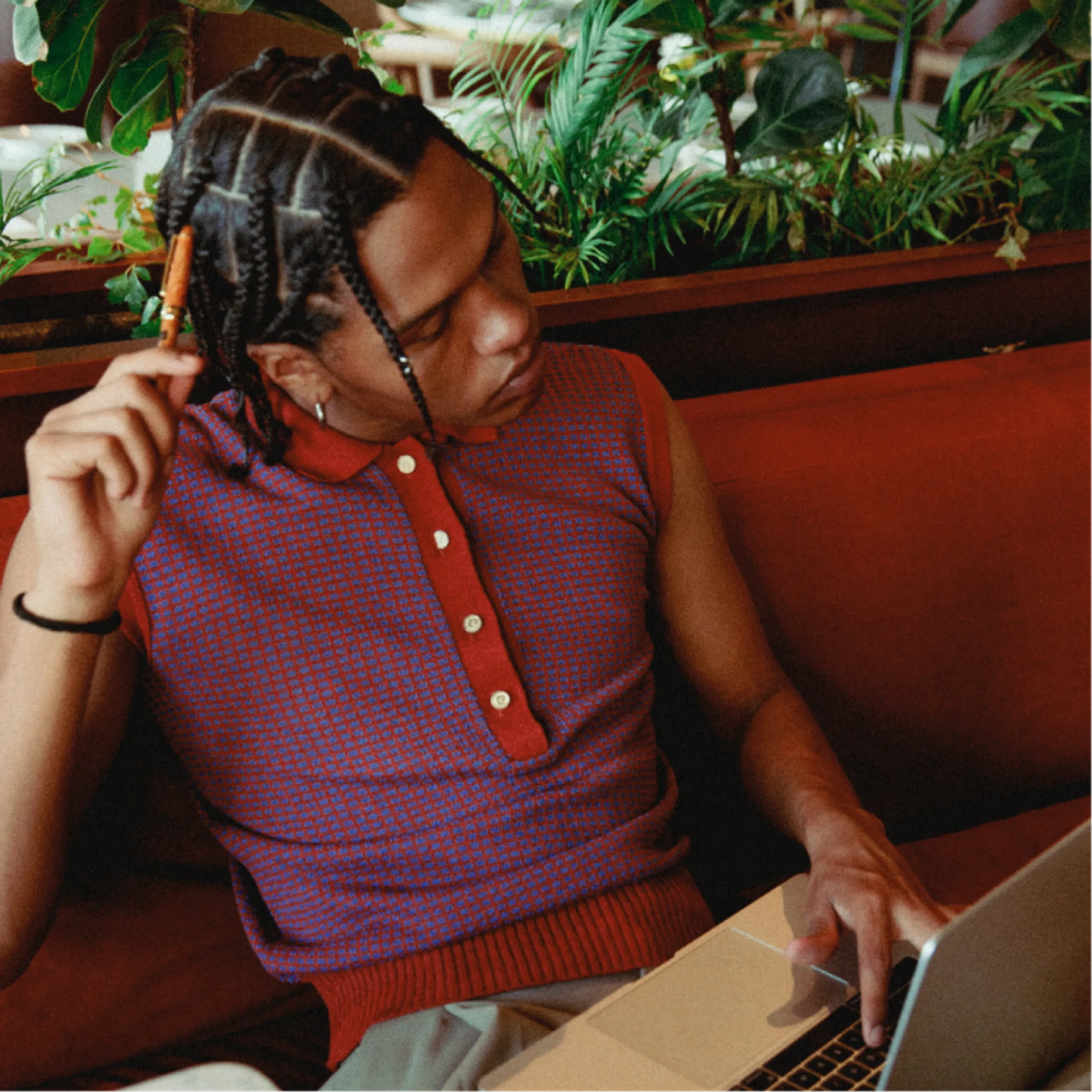 A person with braided hair in a sleeveless red and blue top sits on a red couch, holding a pen and looking at a laptop, surrounded by green plants.
