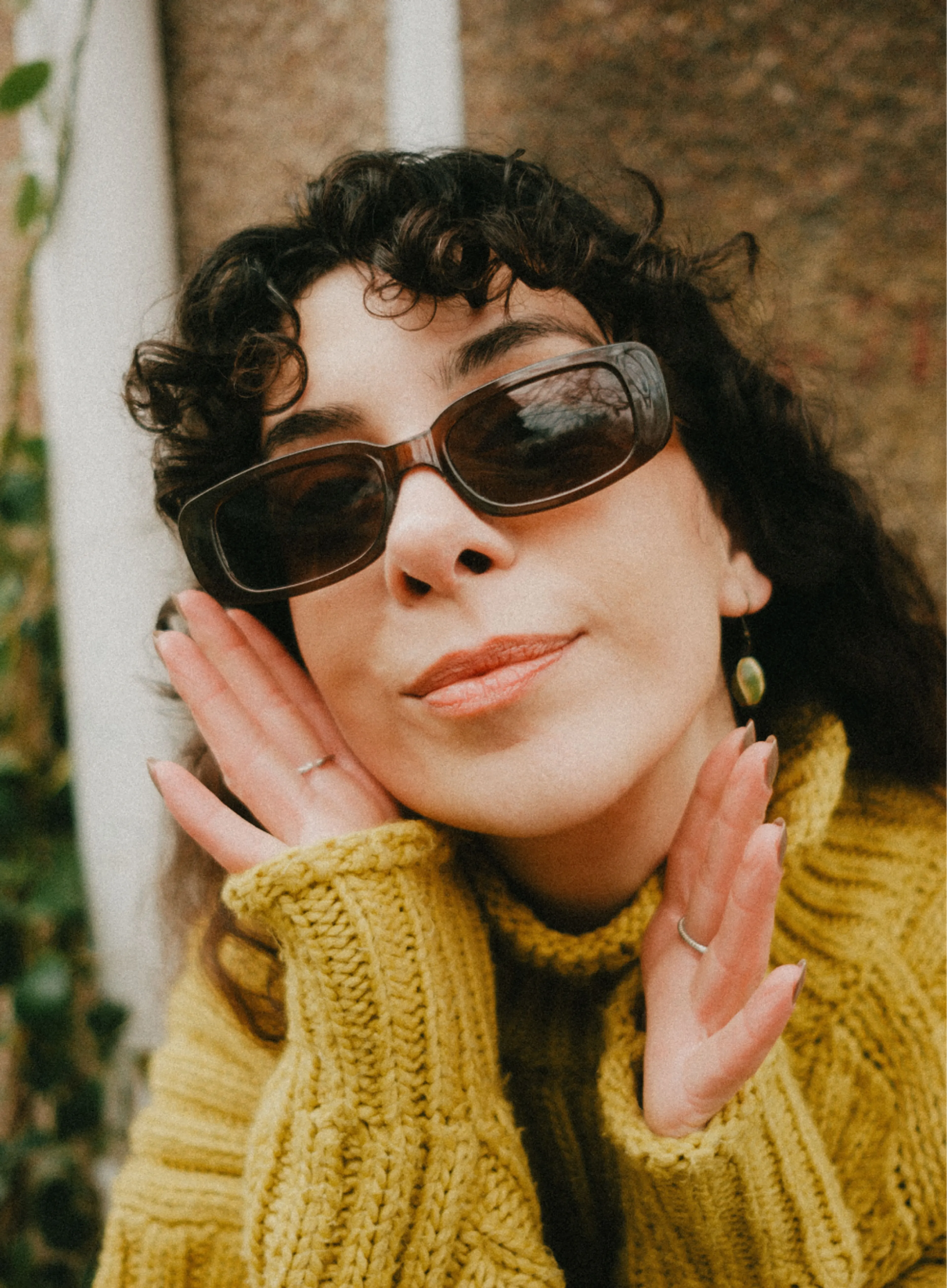 Woman with curly dark hair wearing sunglasses and a chunky yellow sweater, posing with her hands framing her face and smiling slightly. The background is textured with some greenery visible.