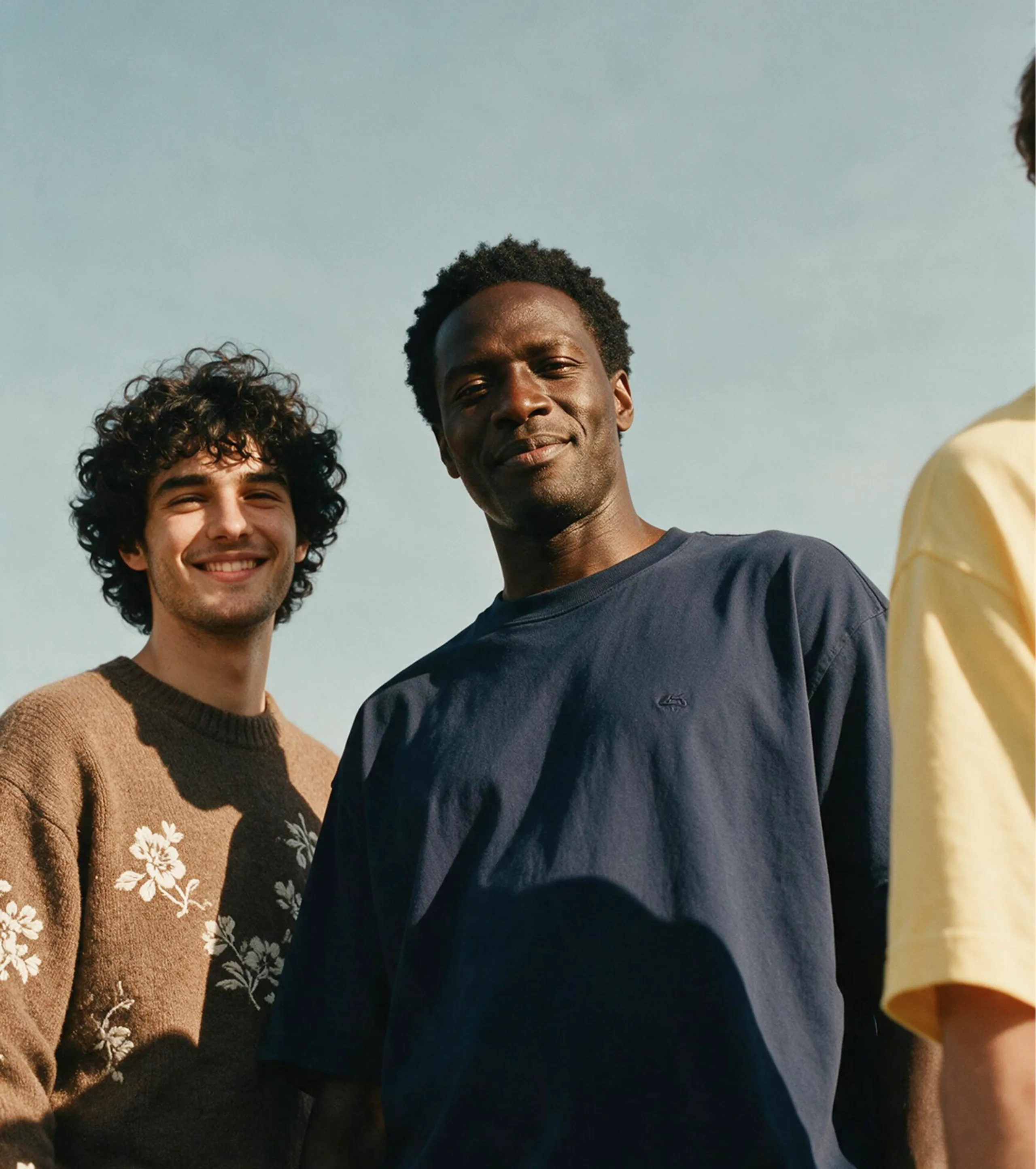 Three people stand outside under a blue sky. Two are clearly visible: one smiling and wearing a brown sweater with white floral patterns, and the other in a navy shirt, also smiling. The third wears yellow and is partly out of frame.