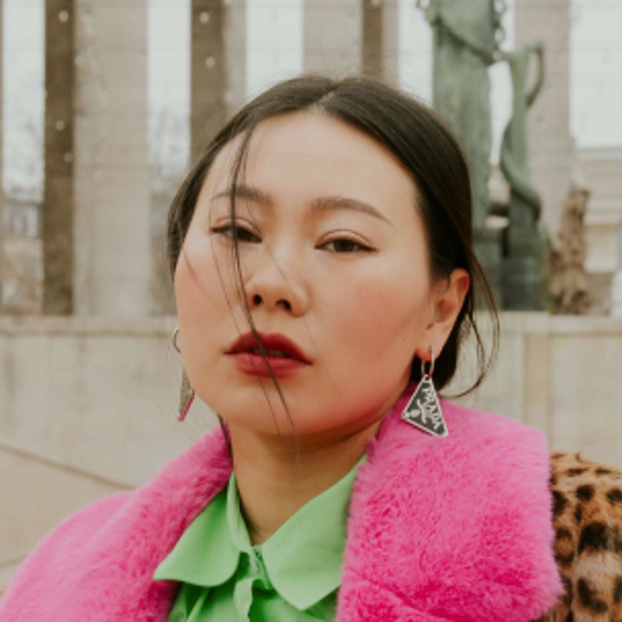 Woman with dark hair wears bold red lipstick, a neon green shirt, a pink faux-fur coat, and geometric earrings. She stands outdoors with blurred statues and columns in the background.