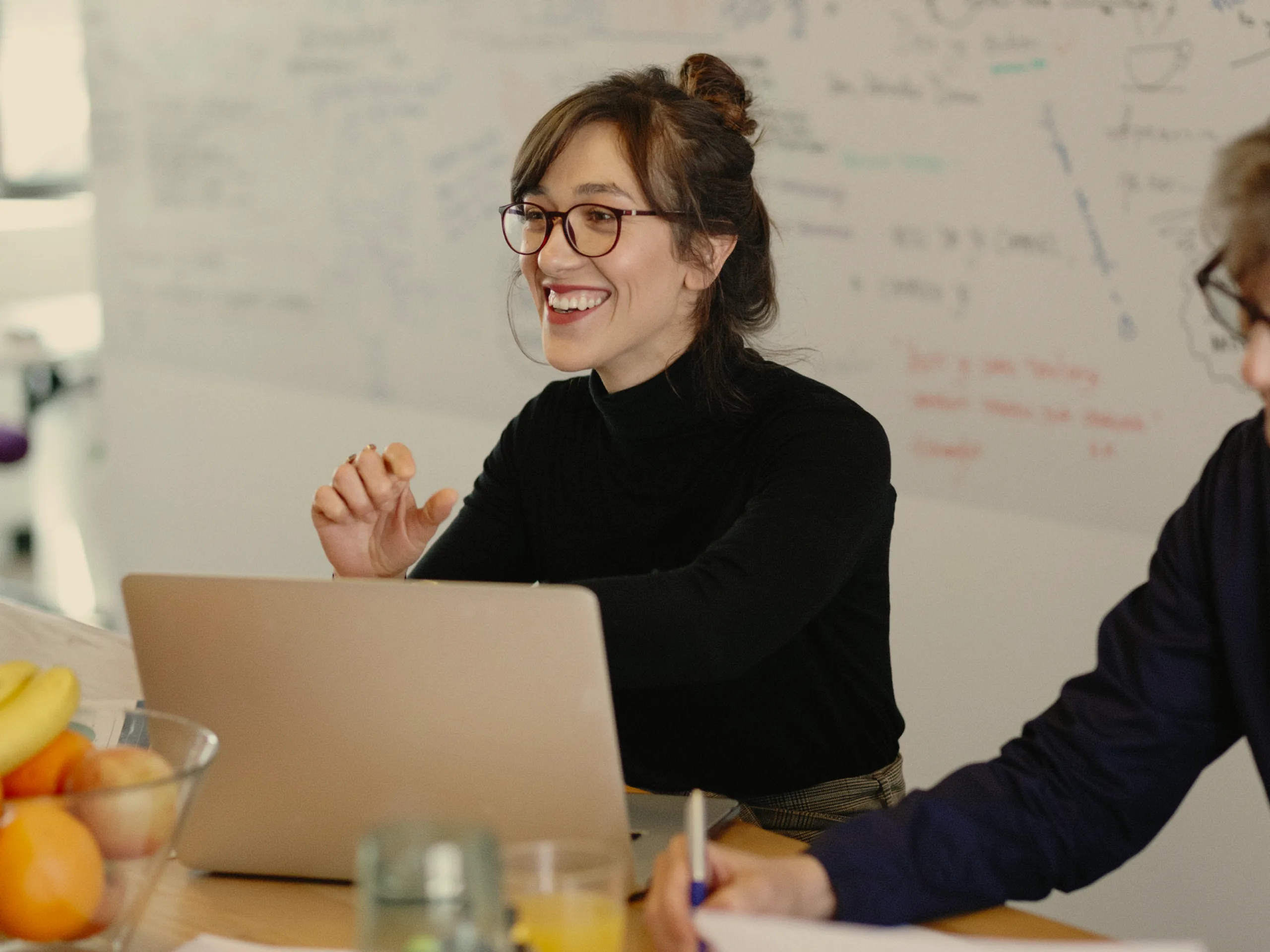A woman wearing glasses and a black top smiles while sitting at a table with a laptop. There is a bowl of fruit and a whiteboard with notes in the background.