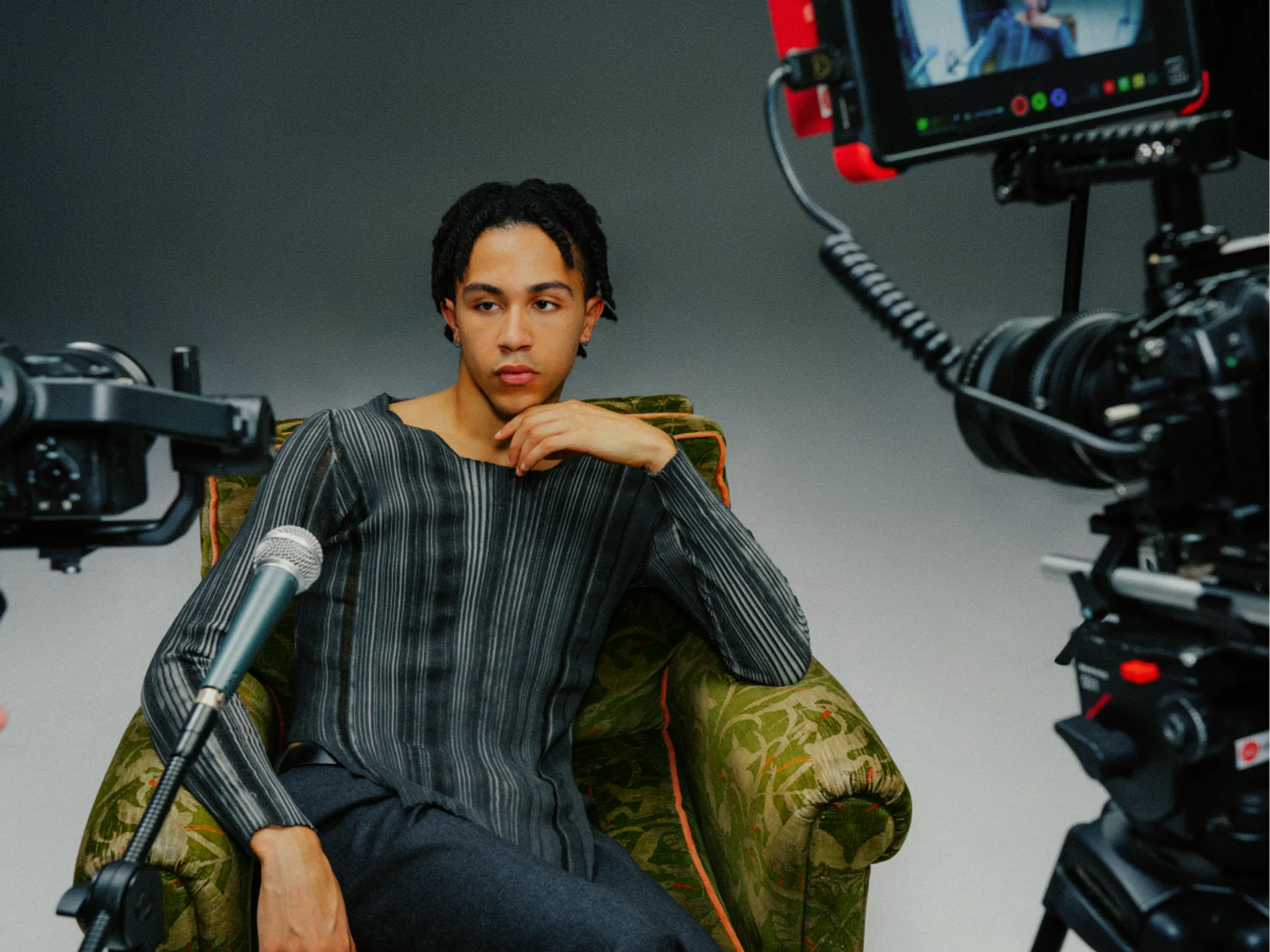 A person sits on a green patterned armchair, posing thoughtfully for an interview while surrounded by cameras and a microphone in a studio setting.