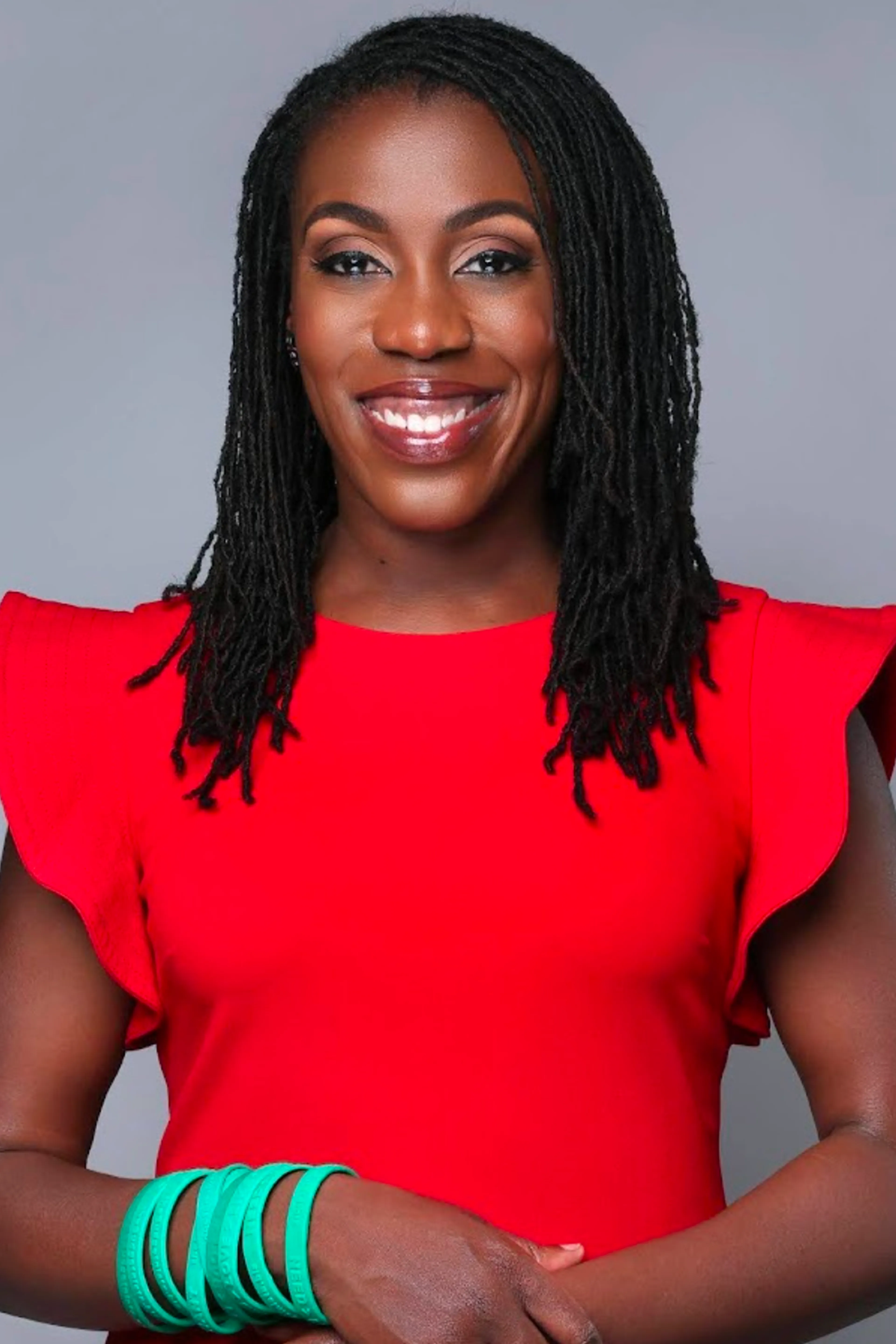 A woman with long braided hair wears a bright red dress with ruffled sleeves and stacked green bangles, smiling against a plain gray background.