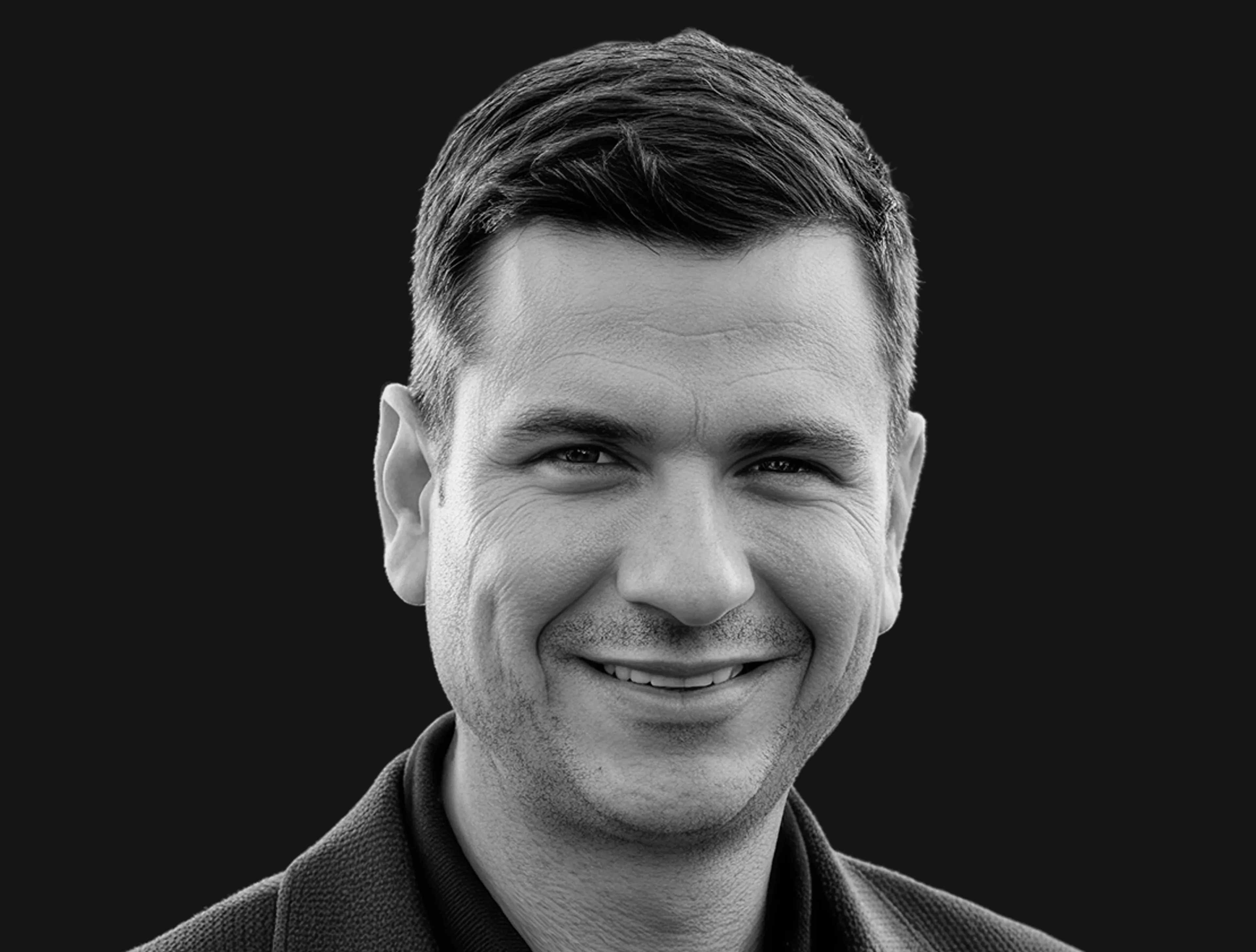A man with short dark hair and a slight smile, wearing a dark collared shirt or jacket, photographed against a solid black background.