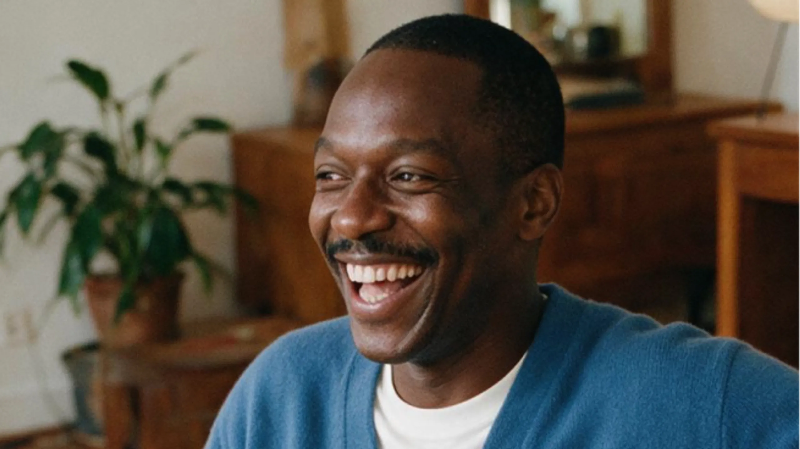 A man wearing a blue sweater smiles and laughs while sitting indoors, with a leafy green plant and wooden furniture in the background.