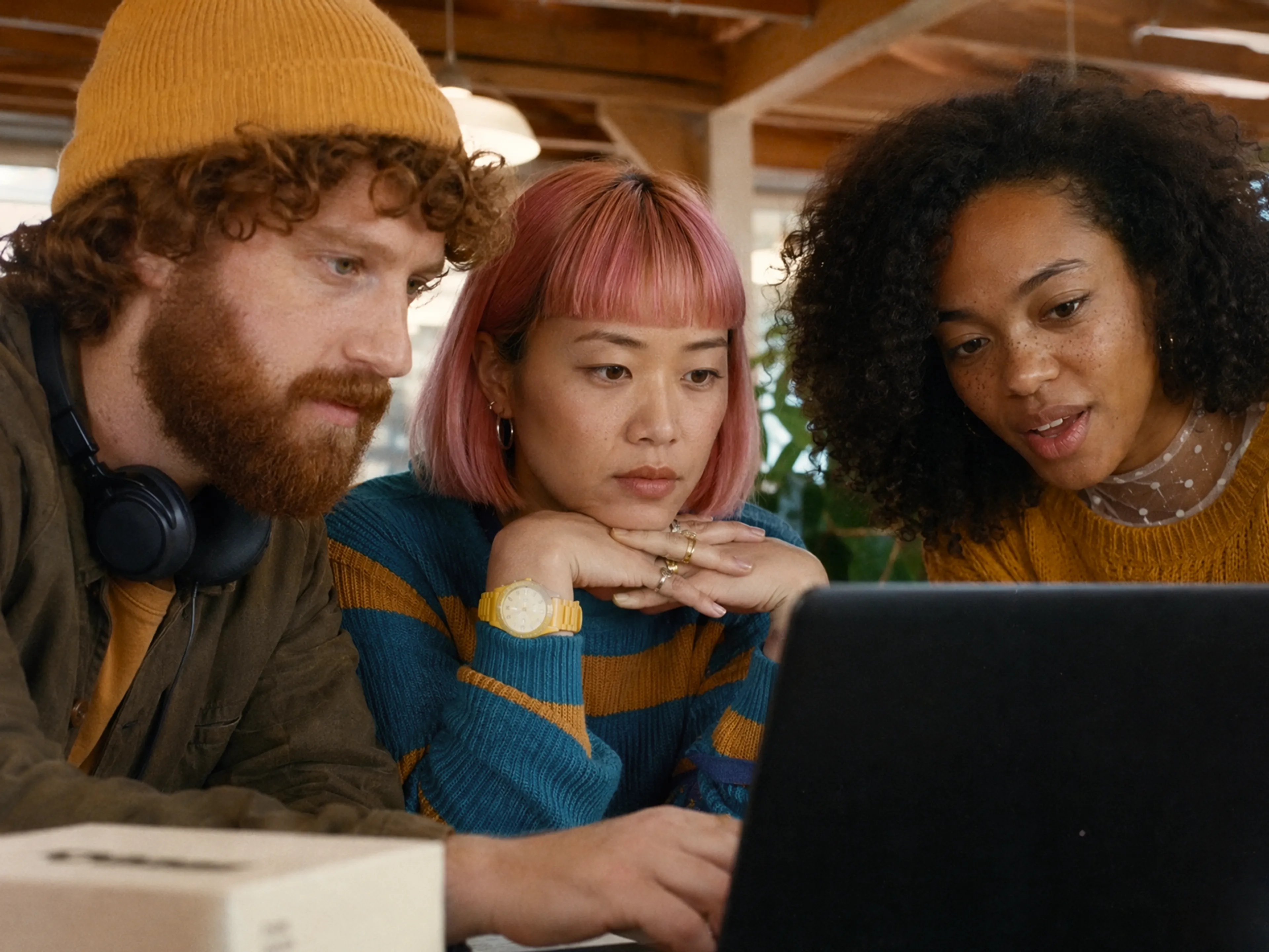 Three young adults, two women and one man, sit closely together, focused on a laptop screen in a cozy indoor setting. They appear to be collaborating or discussing something on the computer.
