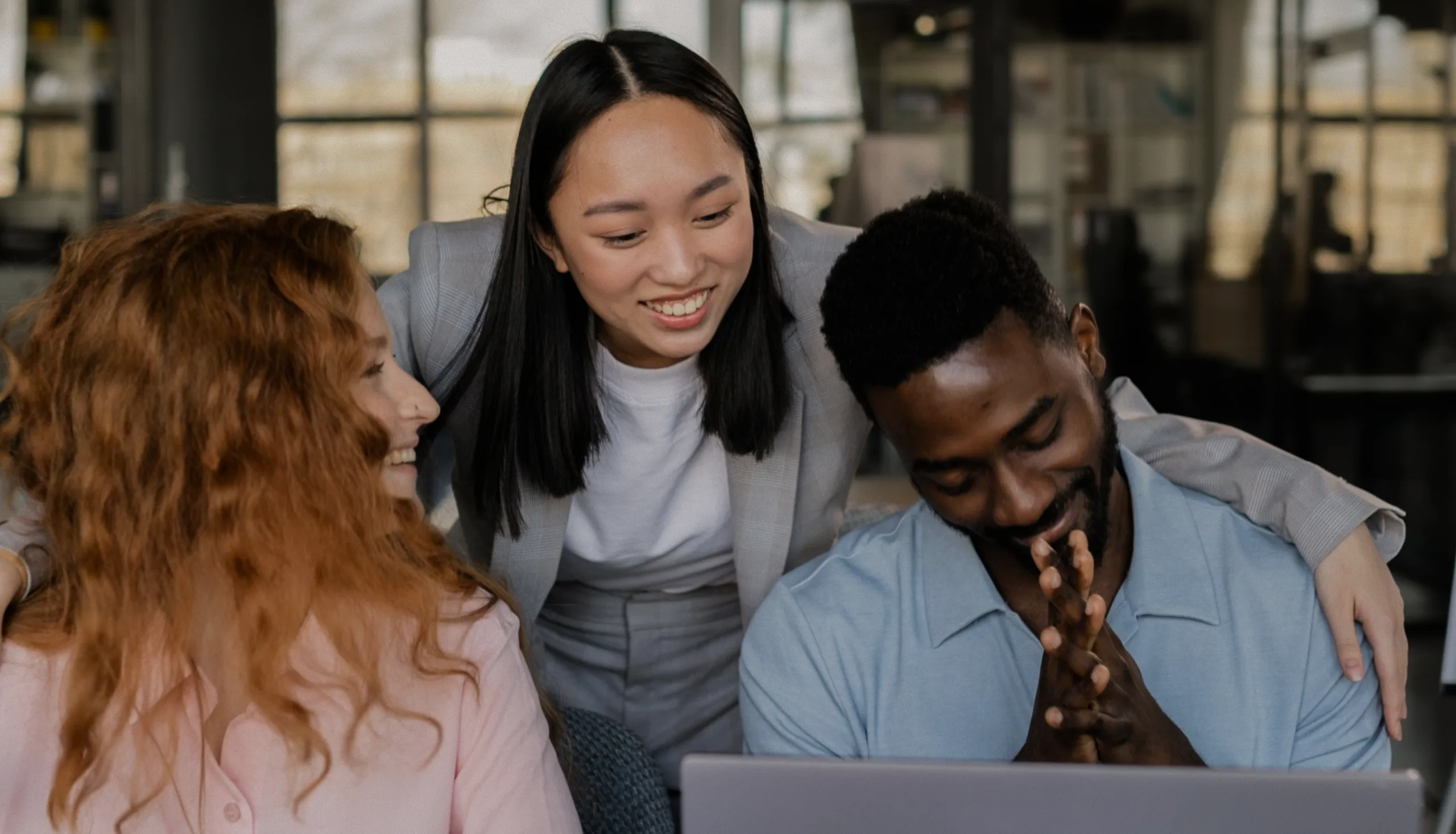 Three colleagues smile and interact around a laptop in an office setting. One person stands, placing arms around the shoulders of the two seated coworkers, all appearing happy and engaged.